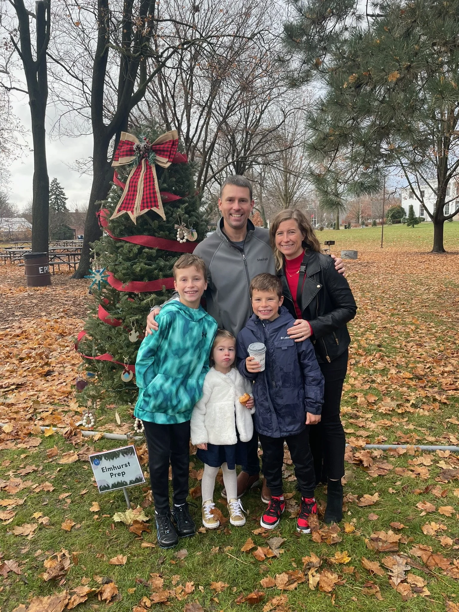 A group of five people, including children and adults, standing outdoors near a decorated Christmas tree in a park.