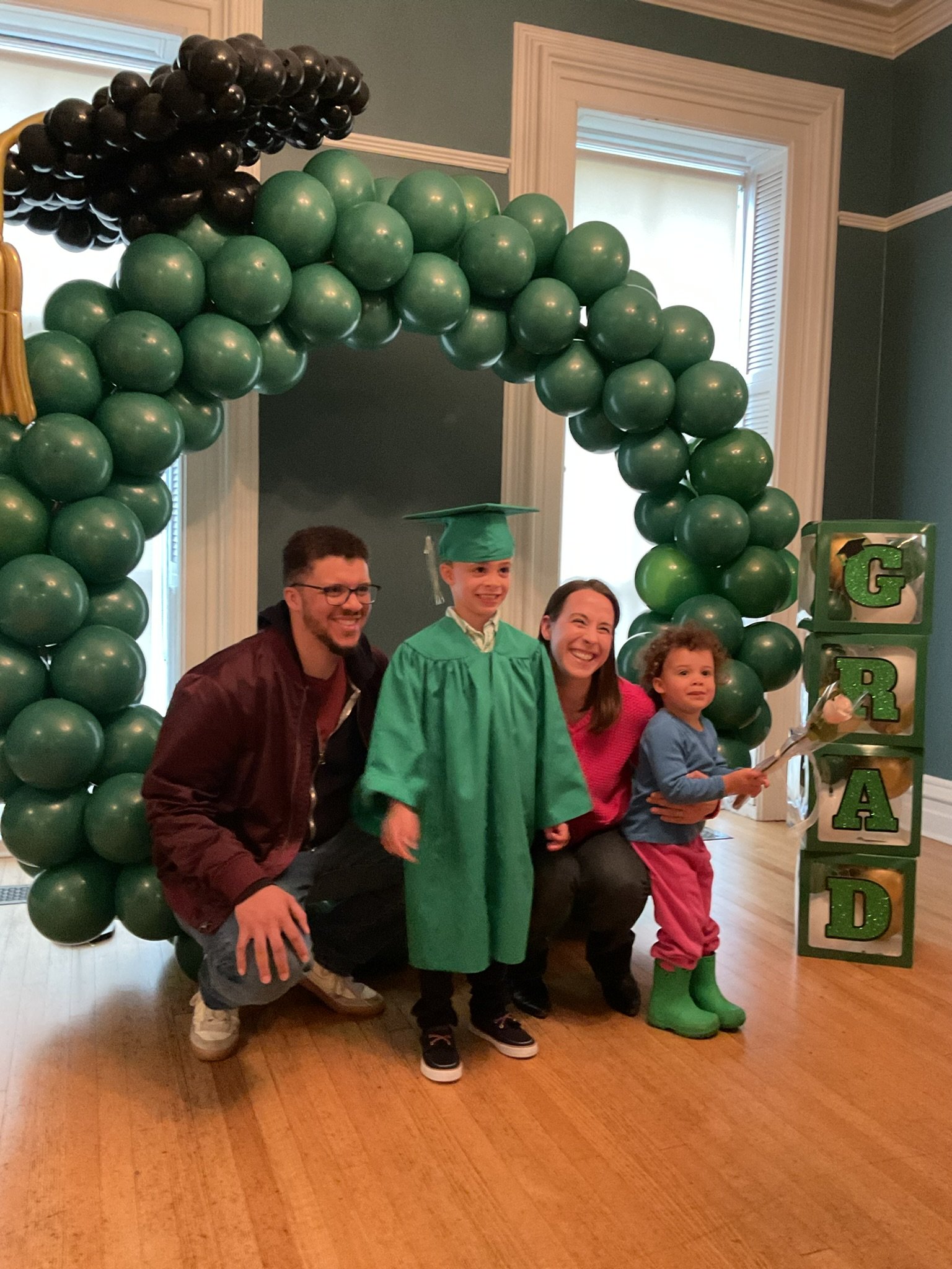 Group of five people, including a child in a green graduation cap and gown, smiling and posing indoors with balloons and a decorative block sign that spells 'GRAD'.