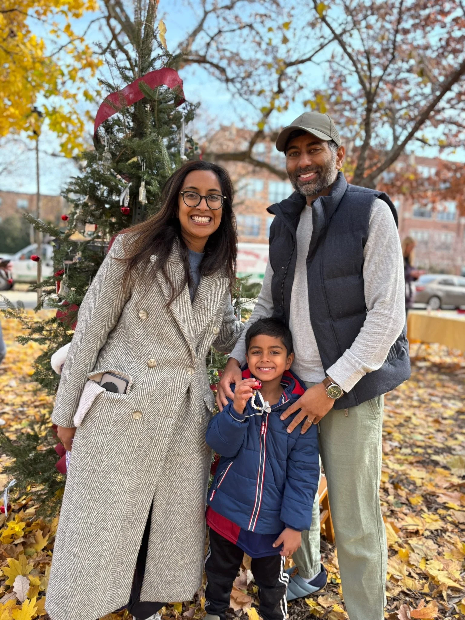 A smiling family of three standing outdoors in front of a Christmas tree.