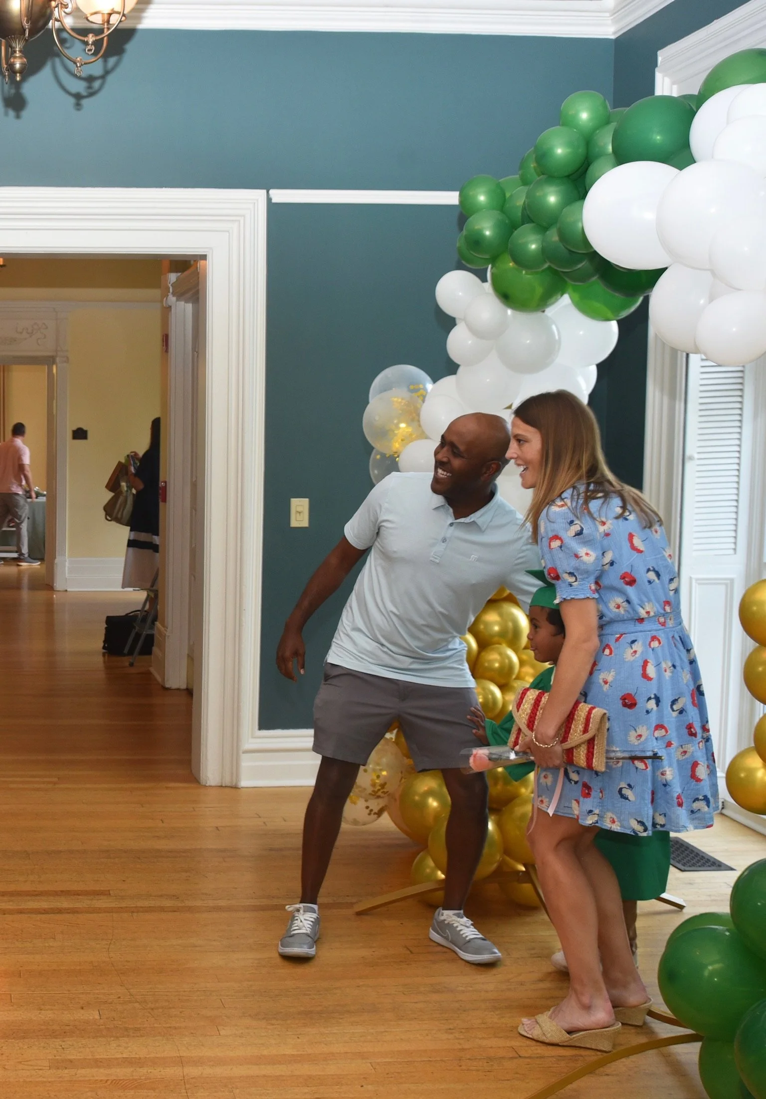 A man and a woman smiling and talking in front of a balloon decoration with green, gold, and white balloons. The woman is holding a woven clutch and wearing a blue dress with cartoon characters, and the man is wearing a light-colored polo shirt and s