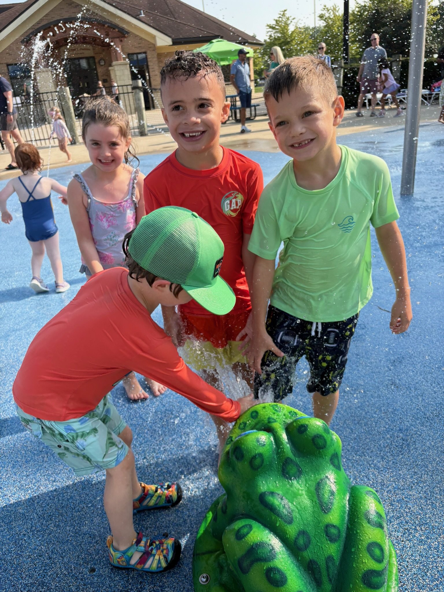 Three children playing on a splash pad, trying to break a green dinosaur toy with water, with others and adults in the background on a sunny day at a recreational area.