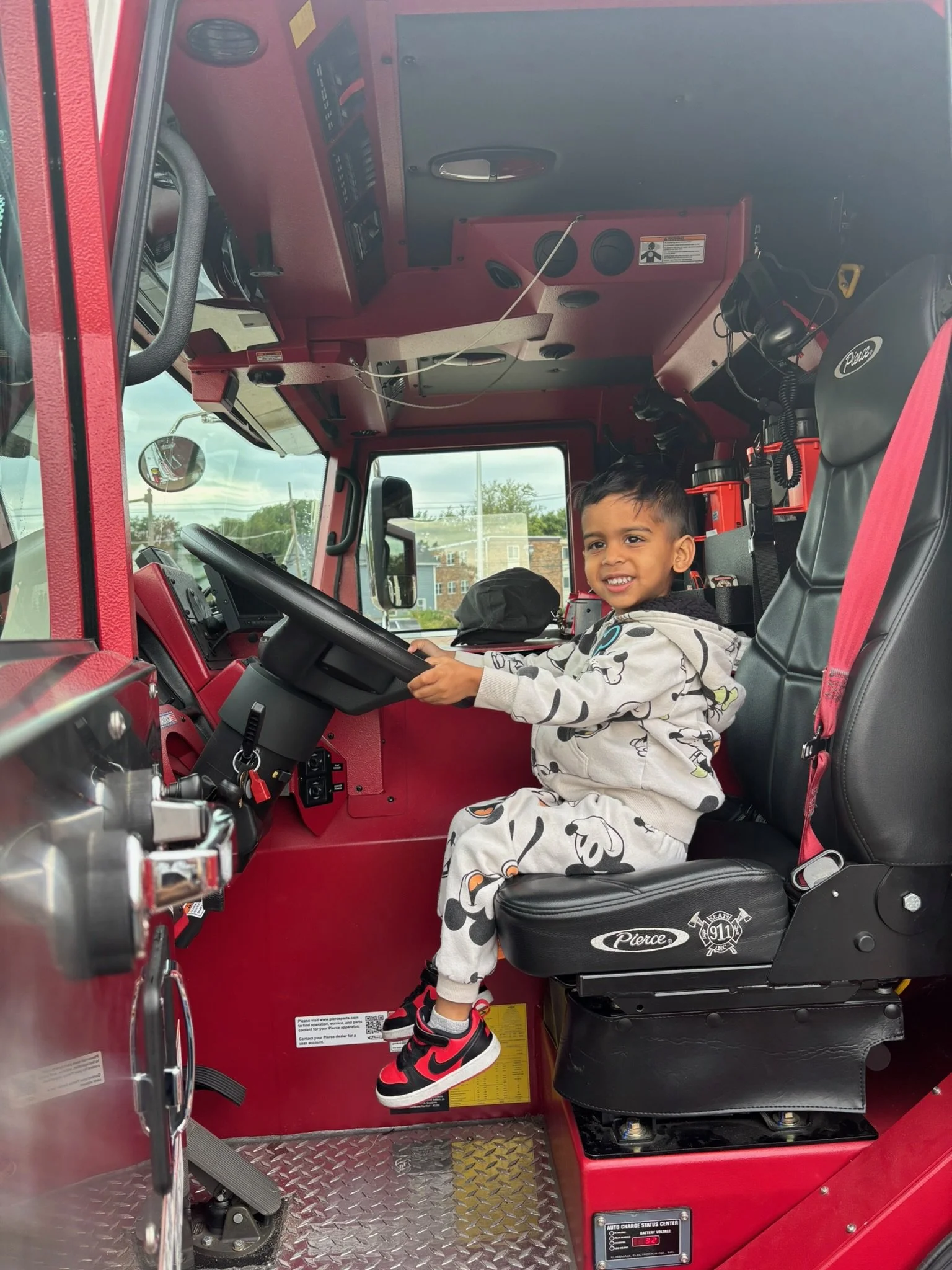 A young boy in pajamas with black, white, and red sneakers sits in the driver's seat of a fire truck, smiling.