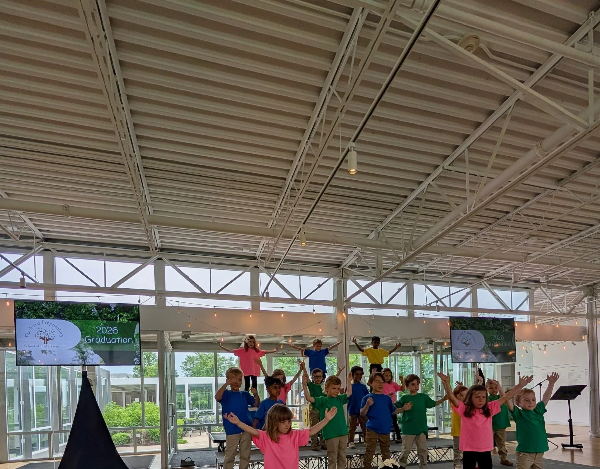 Children performing on a stage at a graduation event inside a building with large windows, displaying banners for 2026 graduation at Elmhurst Prekindergarten School of Early Learning.