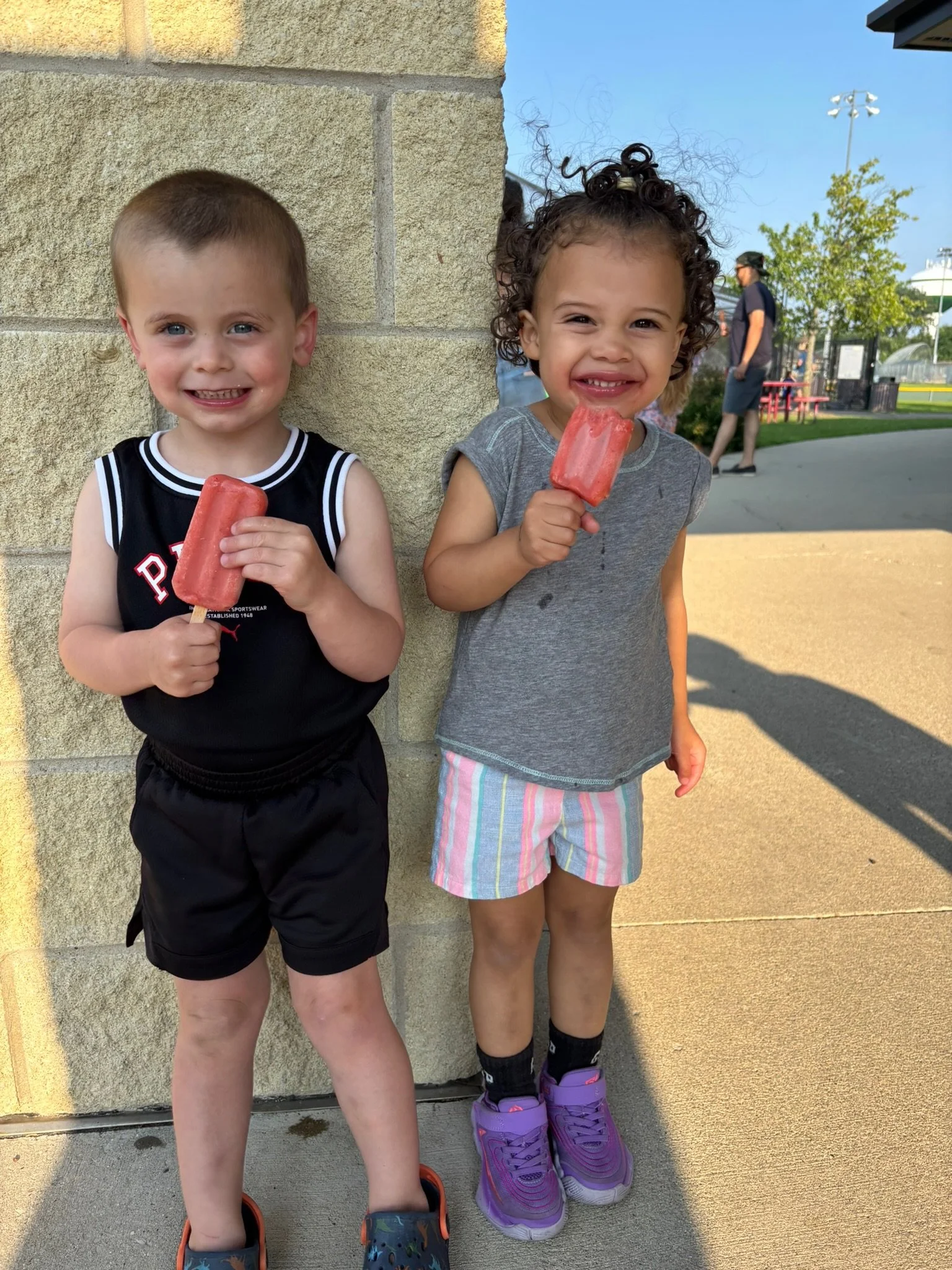 Two young children, a boy and a girl, standing against a stone wall, holding popsicles, smiling at the camera. The boy has short hair, is wearing a black sports jersey and shorts, and colorful Crocs. The girl has curly hair, is wearing a gray t-shirt