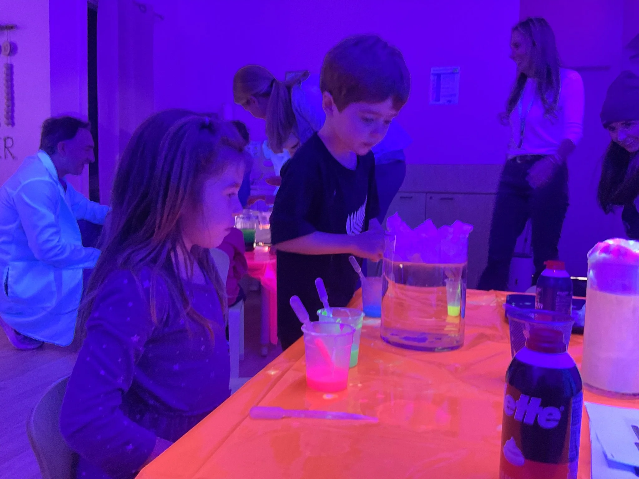 Children and adults gathering around a table with glow-in-the-dark party drinks under blacklight illumination.