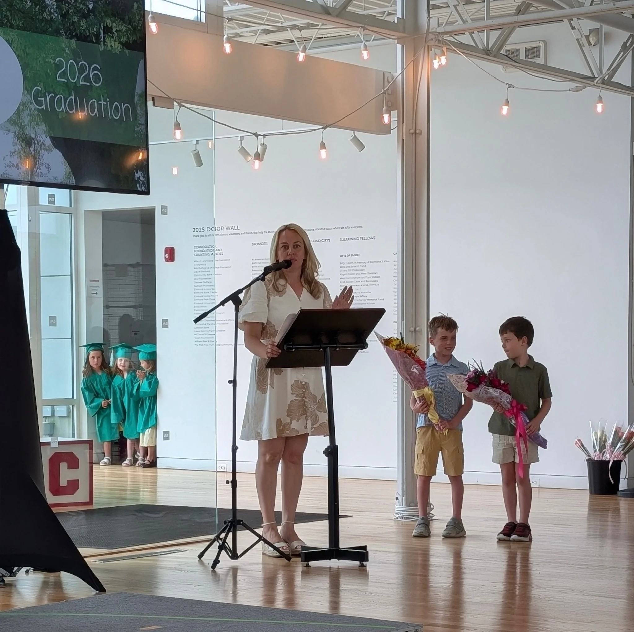 A woman is speaking at a podium with a microphone during a graduation ceremony, holding a bouquet of flowers. Four children in green caps and gowns are visible in the background, and the scene is set in a bright indoor space with large windows, a sig