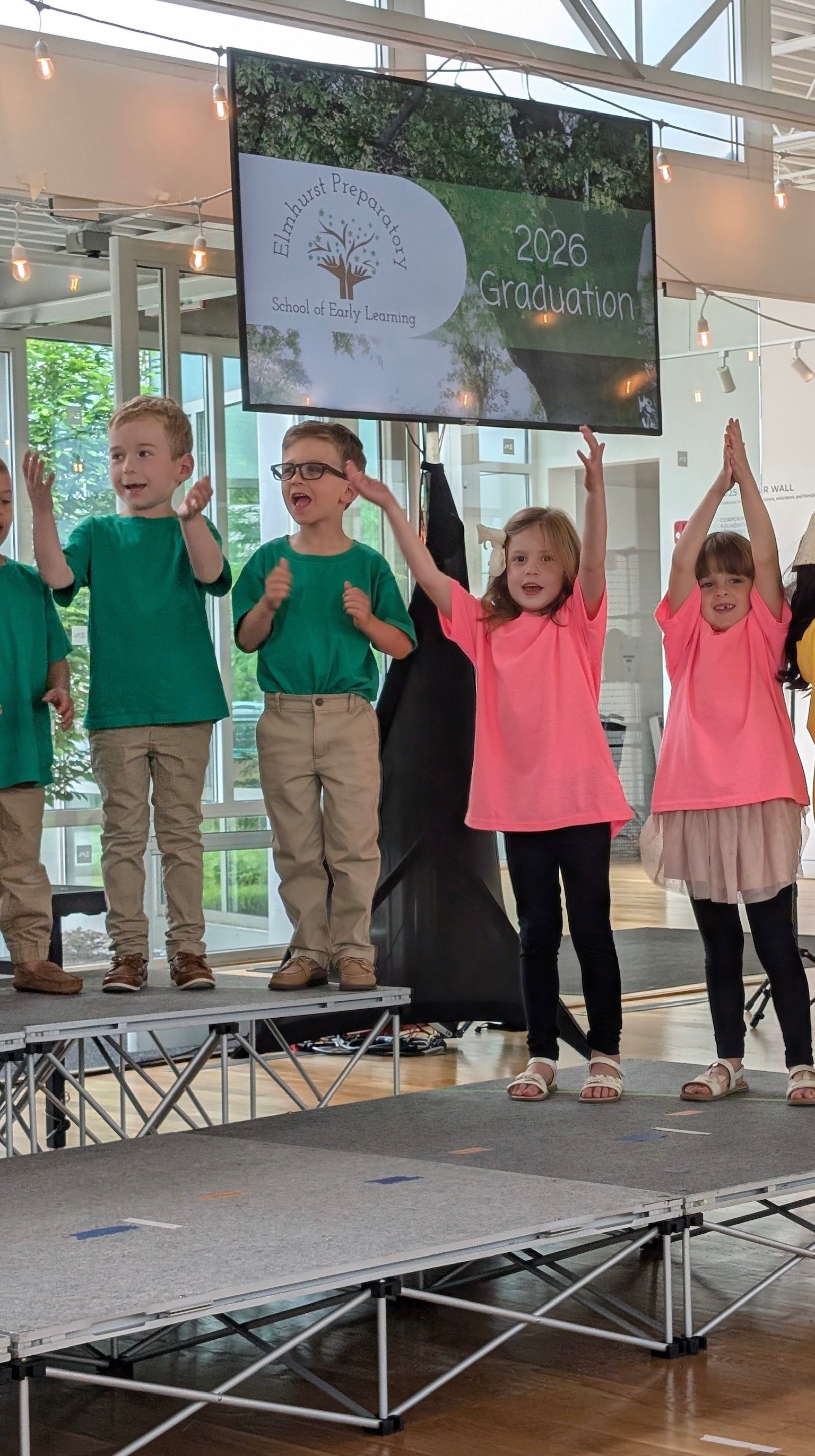 Children on stage at Elmhurst Preparatory School of Early Learning 2026 graduation ceremony, with a large screen displaying the school logo and event details.