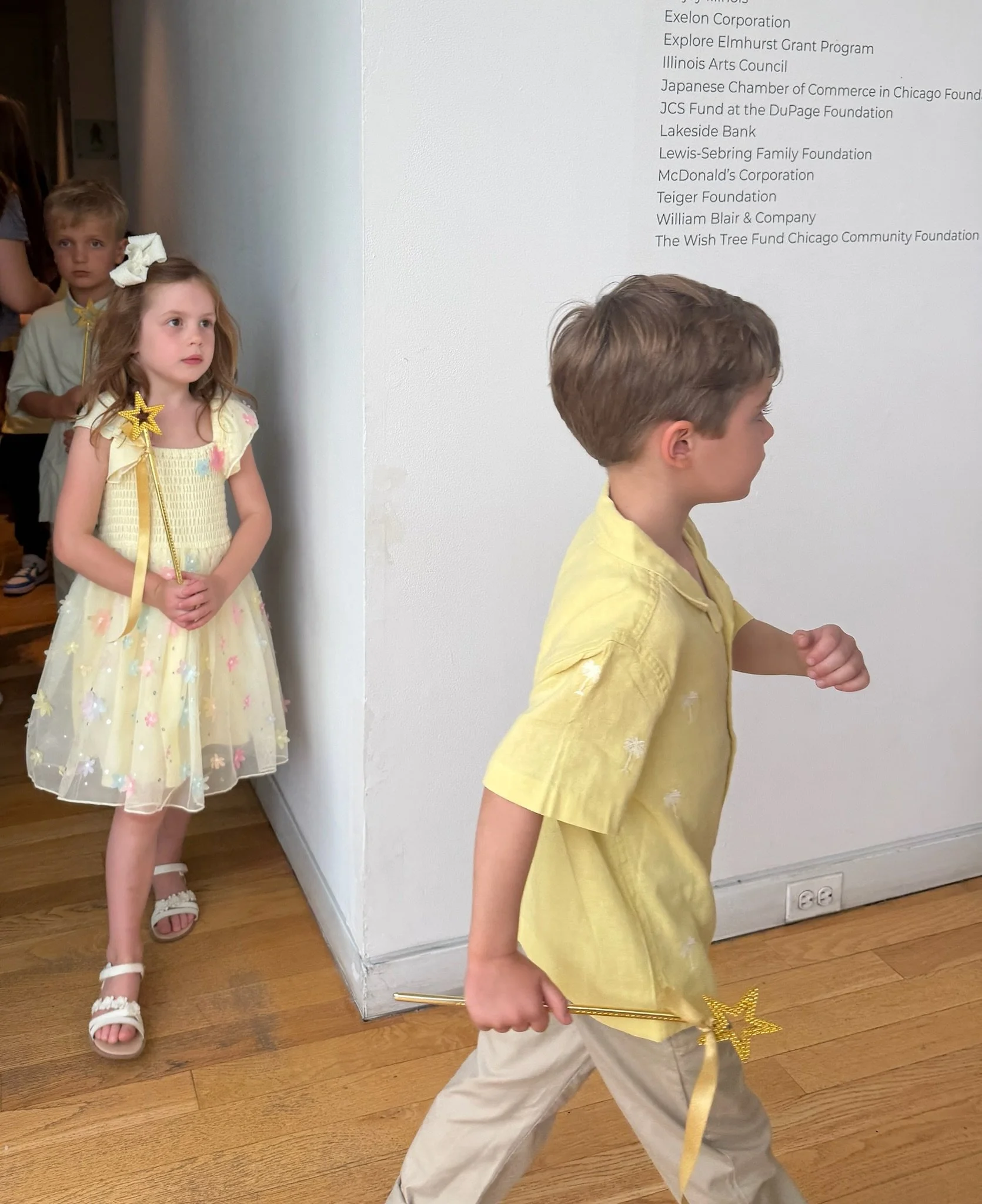 Three children standing indoors, two girls and one boy, with the boy in the foreground and the girls in the background. The girl in front has a yellow dress with pink and blue flower designs and a star wand. The girl behind her has a light yellow dre
