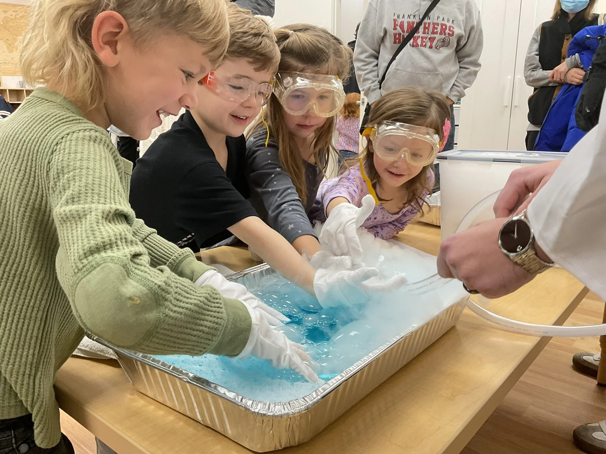 Children wearing safety goggles and gloves working with a blue slime or liquid in a tray on a table, supervised by an adult, in a classroom or science lab setting.