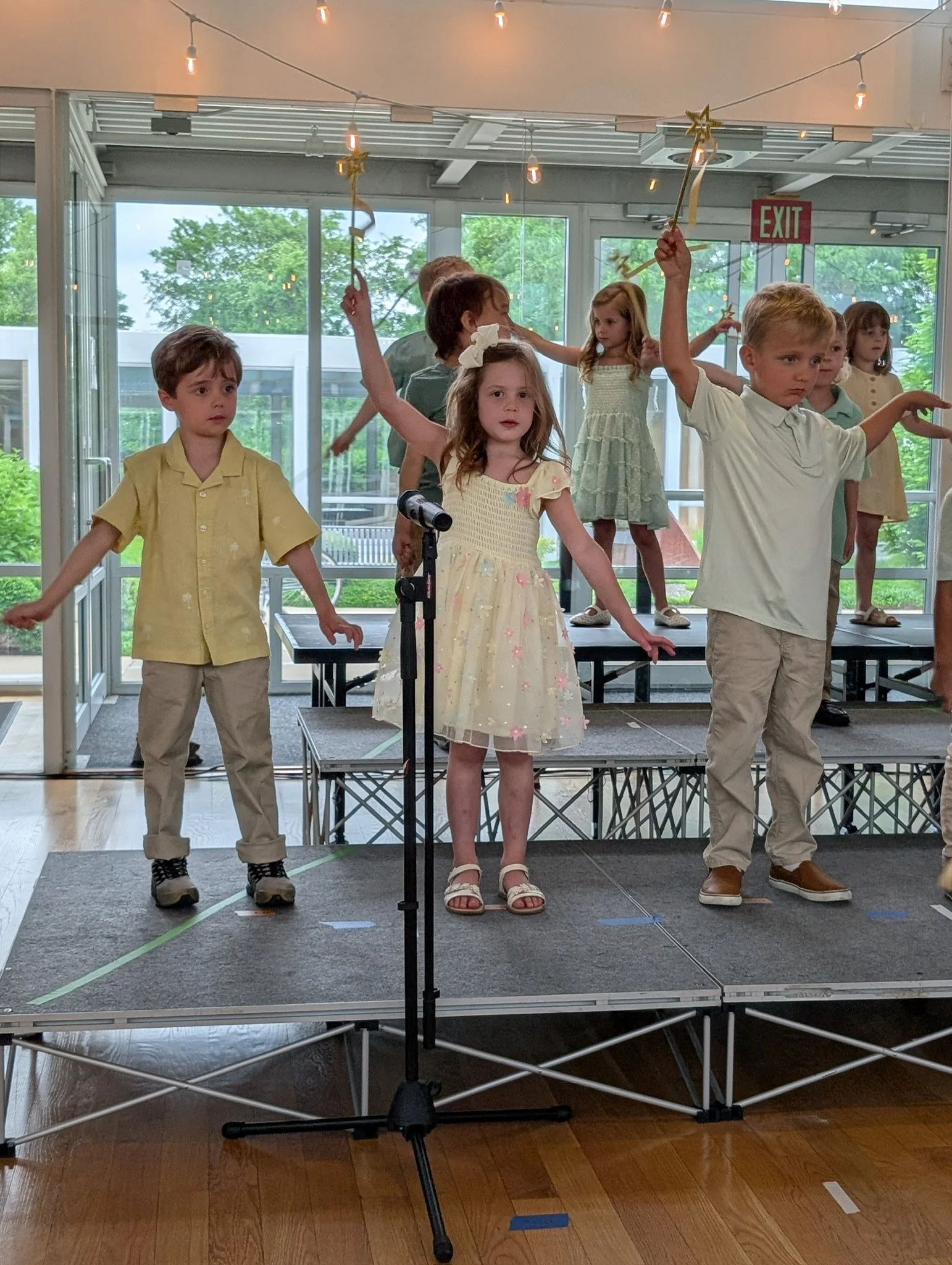 Children performing on stage, some holding stars and wands, with a microphone in front of them.