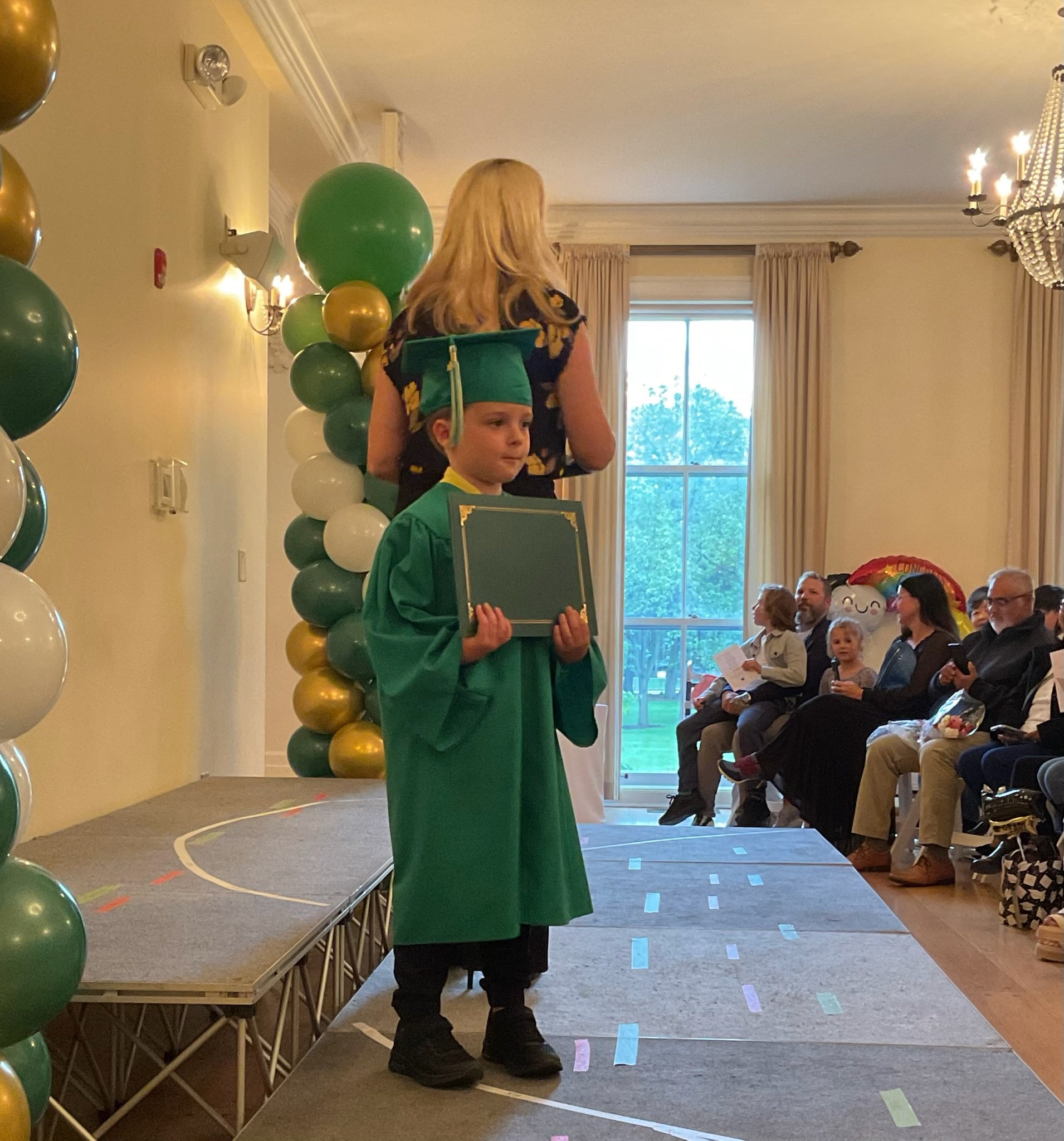 A girl in a black dress with yellow flowers wearing a green graduation cap and gown holding a certificate, standing on a stage decorated with green and gold balloons, with seated audience members and colorful balloons in the background.