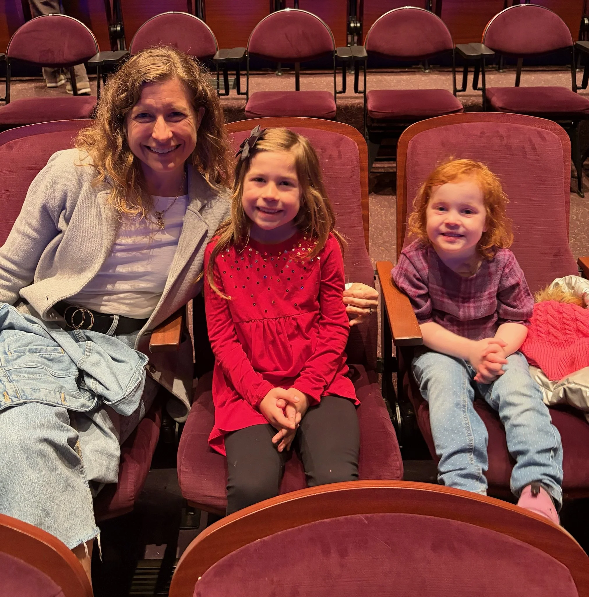 Smiling women and two young girls sitting in burgundy theater seats in an auditorium.