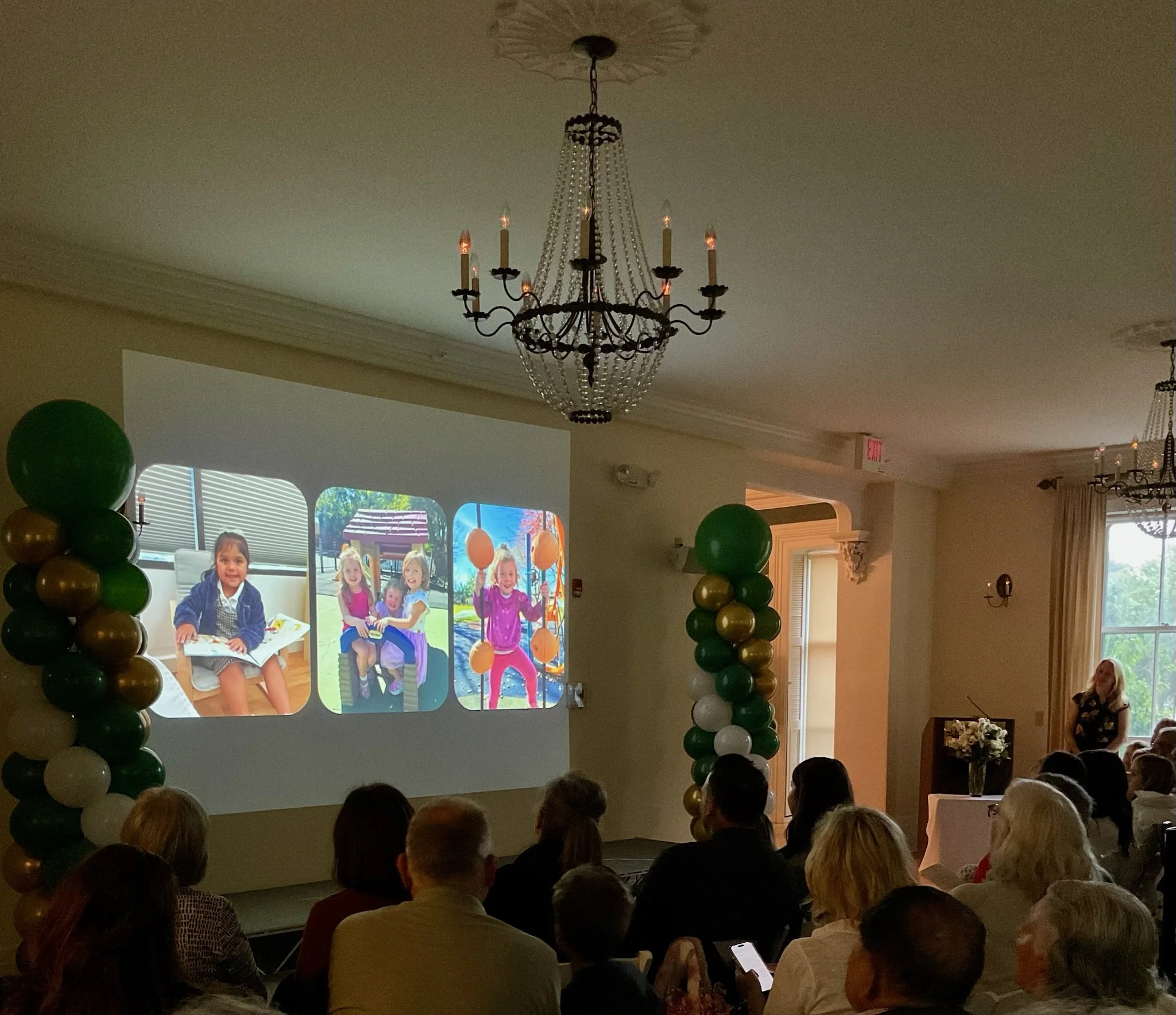 People attending a celebration, with balloon columns on either side of a projection screen displaying children and outdoor scenes, and a chandelier hanging from the ceiling.