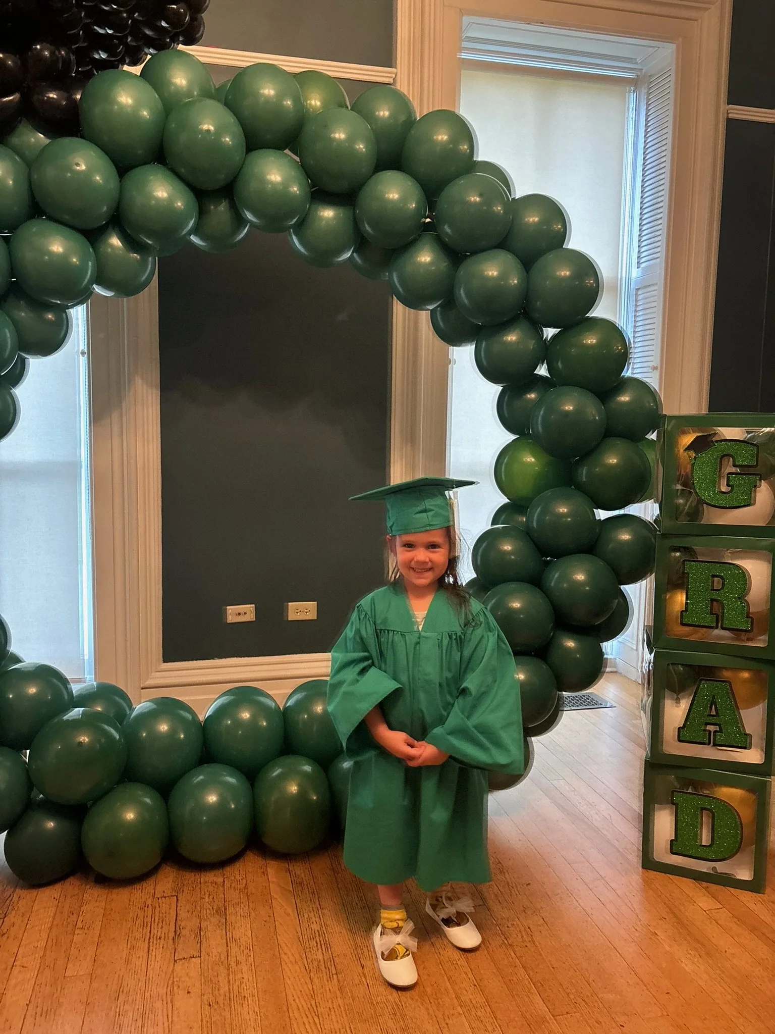 A young girl wearing a green graduation gown and cap, standing in front of a green balloon arch and a sticker display that spells out "GRAD" in glittery green letters.
