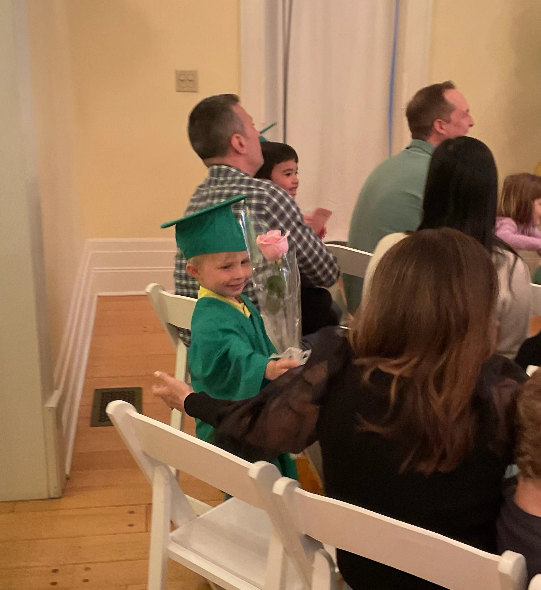 A group of people attending an event, seated in white chairs along a wall. A young boy in a green jacket and graduation cap with a pink flower is in the foreground, smiling. Behind him, a man with a checked shirt and a woman are visible, with the wom