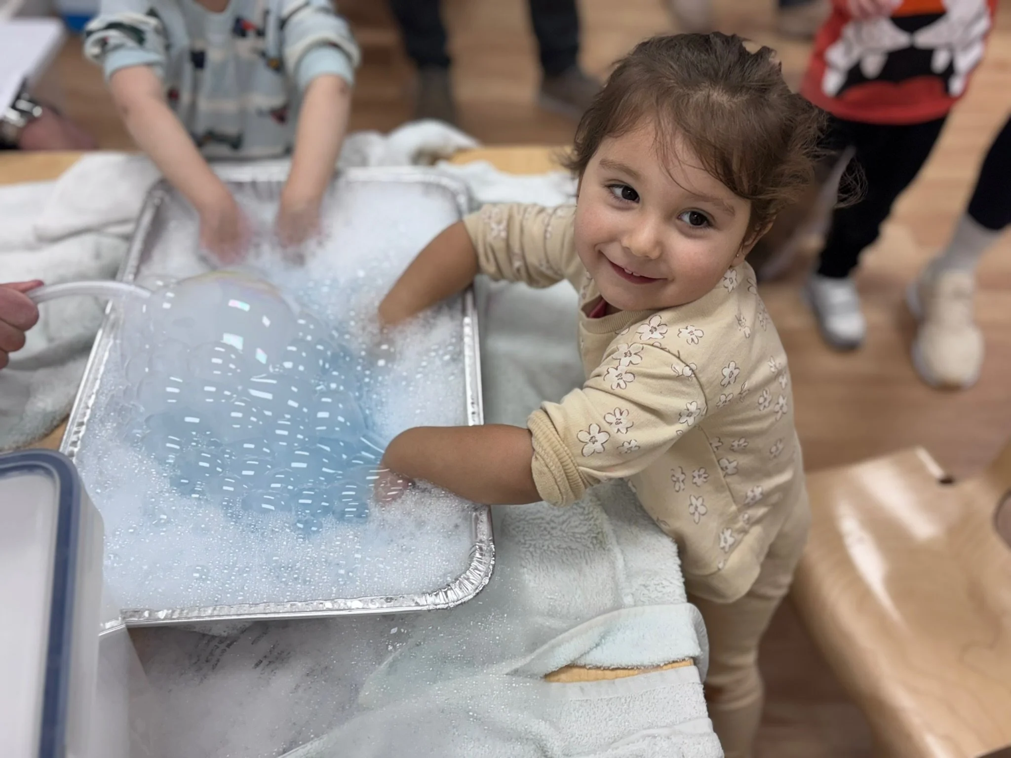 A young child with curly hair washing a bubble-covered blue ball in a tray of soapy water, surrounded by other children at a table.