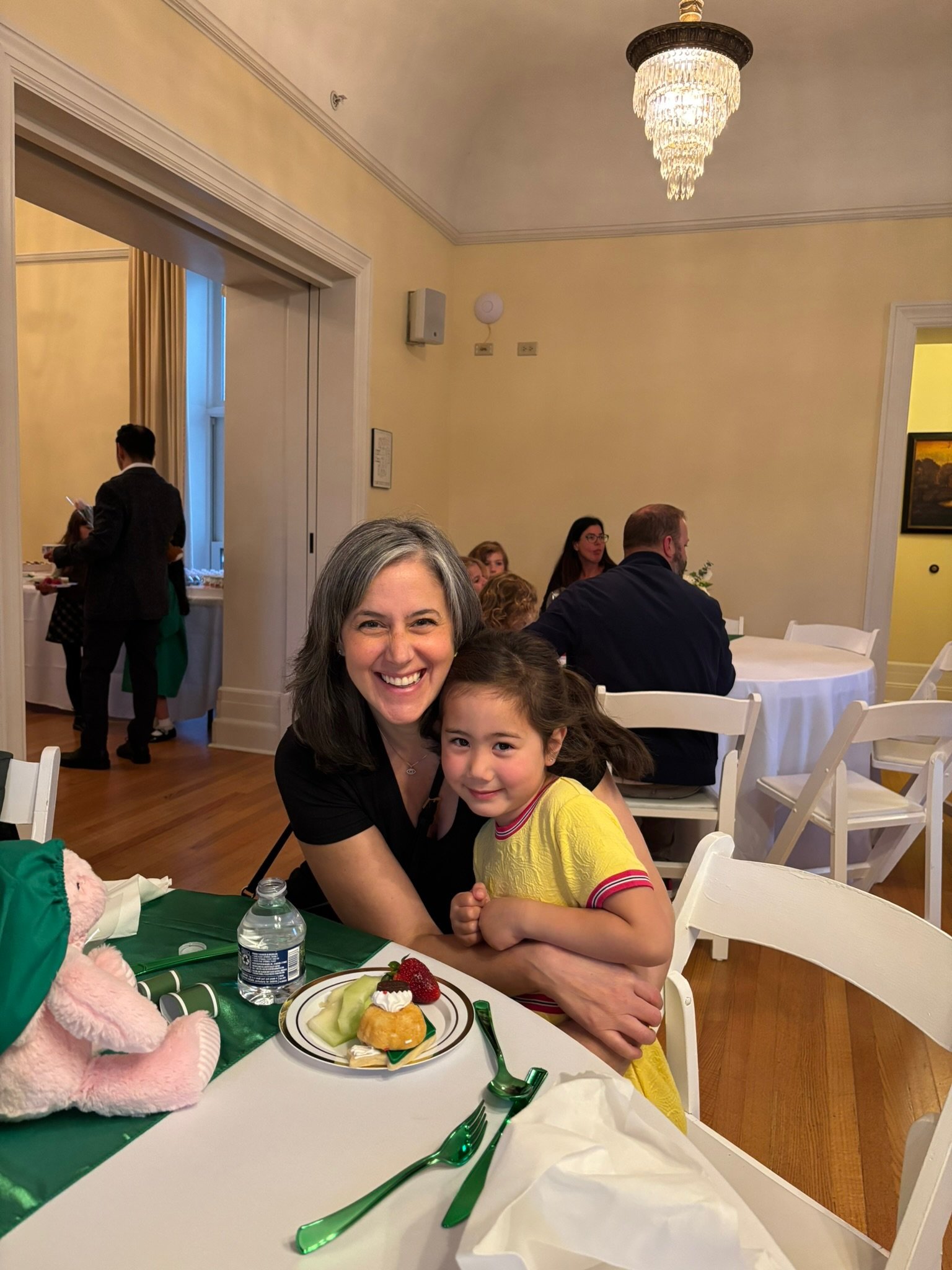 Smiling woman and young girl sitting at a table with dessert, in a well-lit restaurant or banquet hall with other guests in the background.