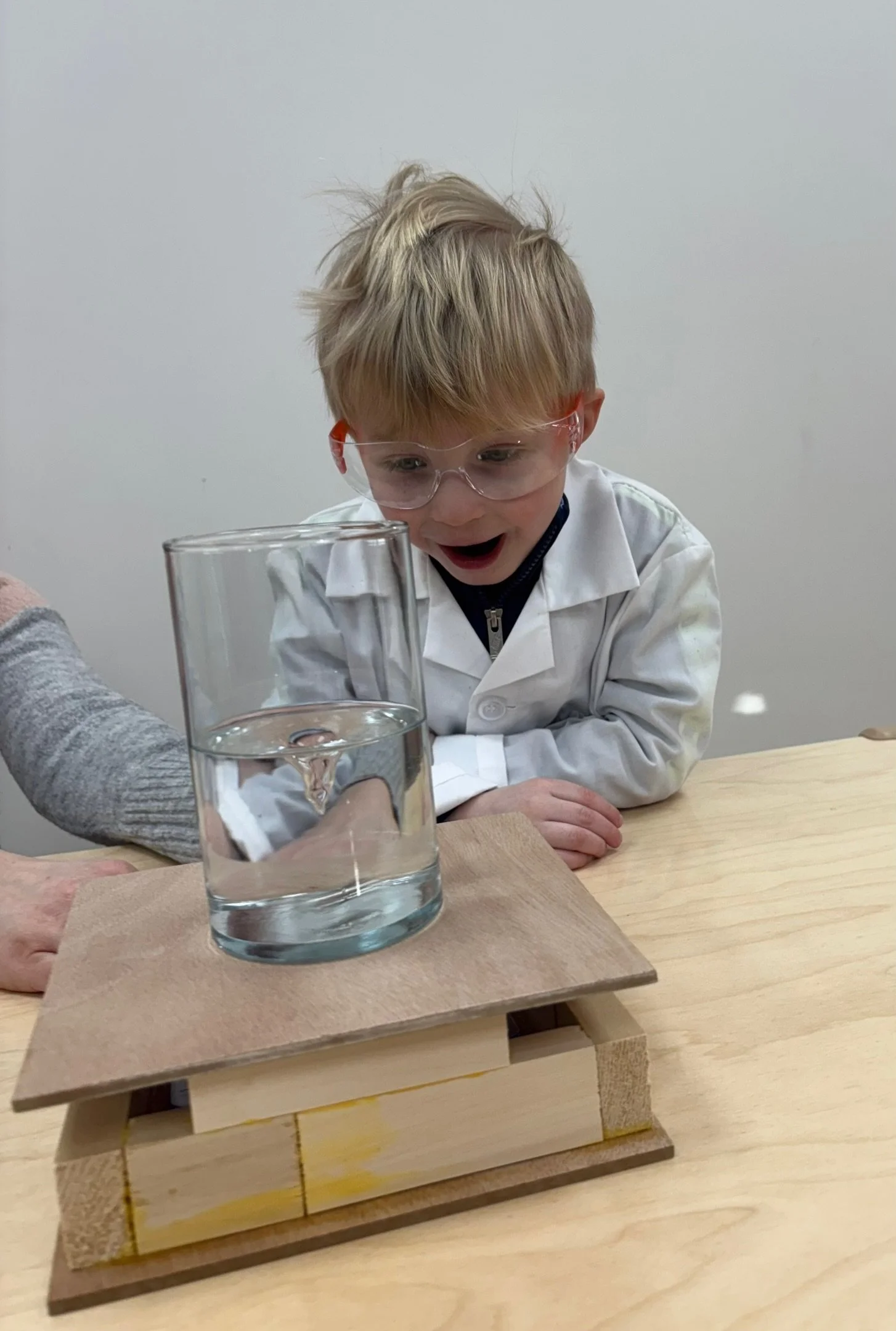 A young boy wearing safety glasses, a white lab coat, sitting at a table, looking excitedly at a glass of water placed on a wooden stand.