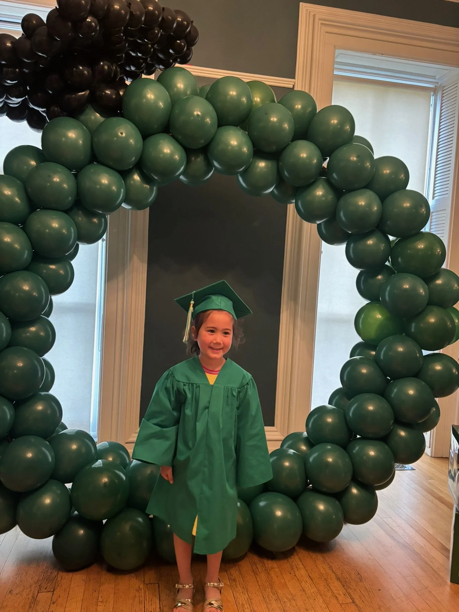 A young girl in a green graduation cap and gown standing in front of a balloon arch made of green and black balloons, celebrating a graduation.