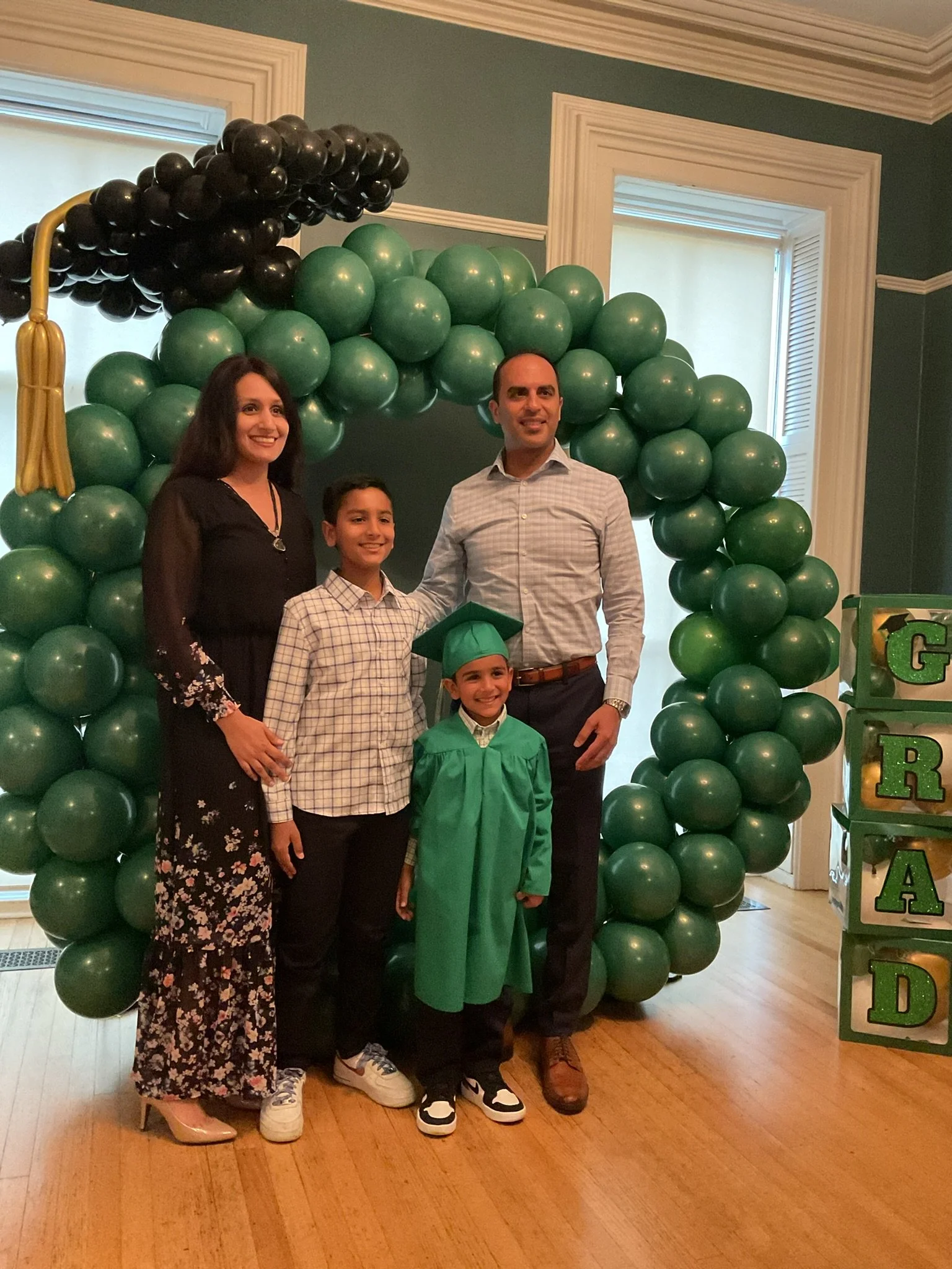 A group of people, including a young boy in a graduation gown and cap, standing inside a room decorated with green and black balloons.