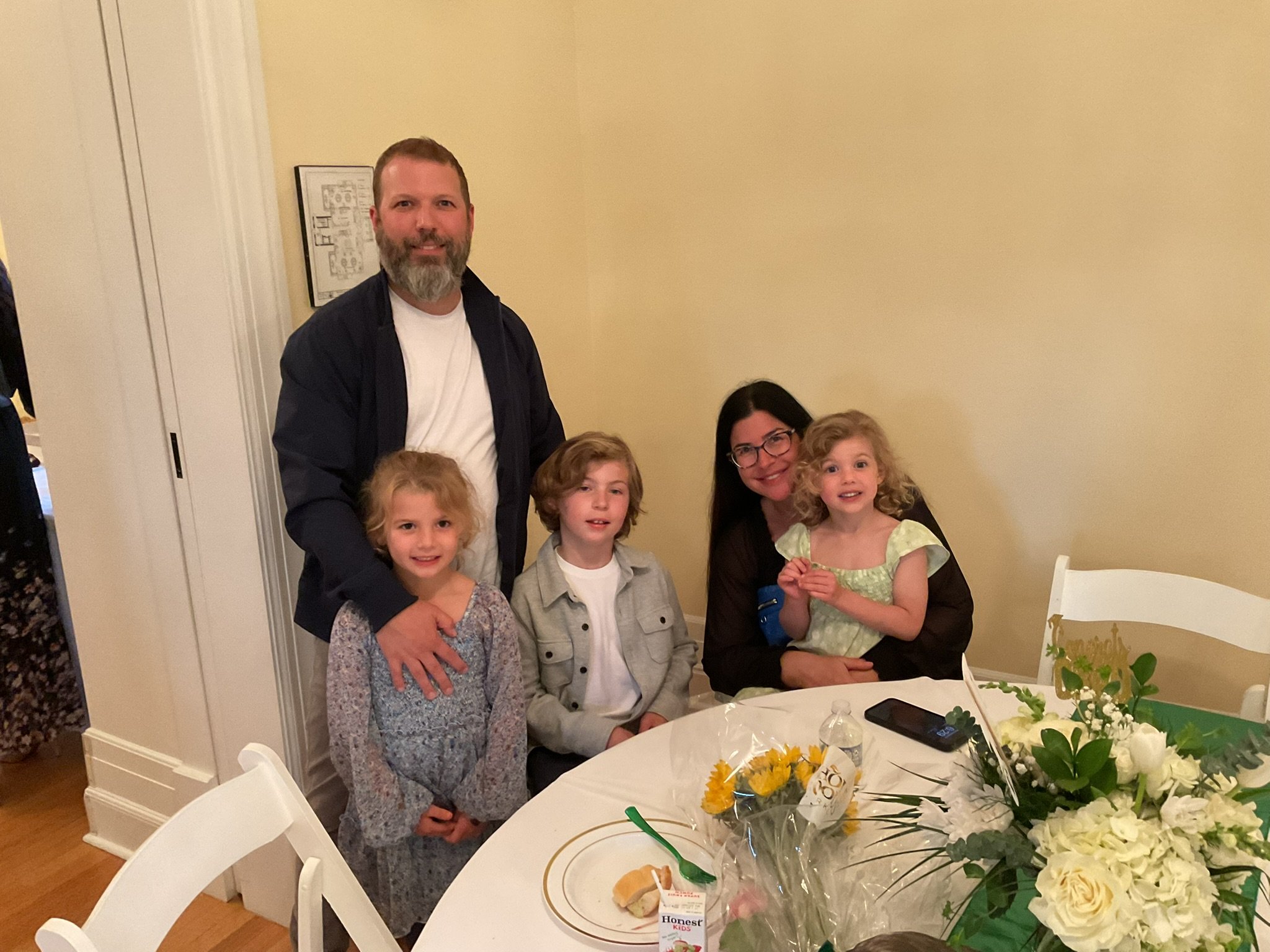 A group of five people, including three children and two adults, gathered around a table during a celebration. The table has floral arrangements and party favors, indicating a festive occasion.