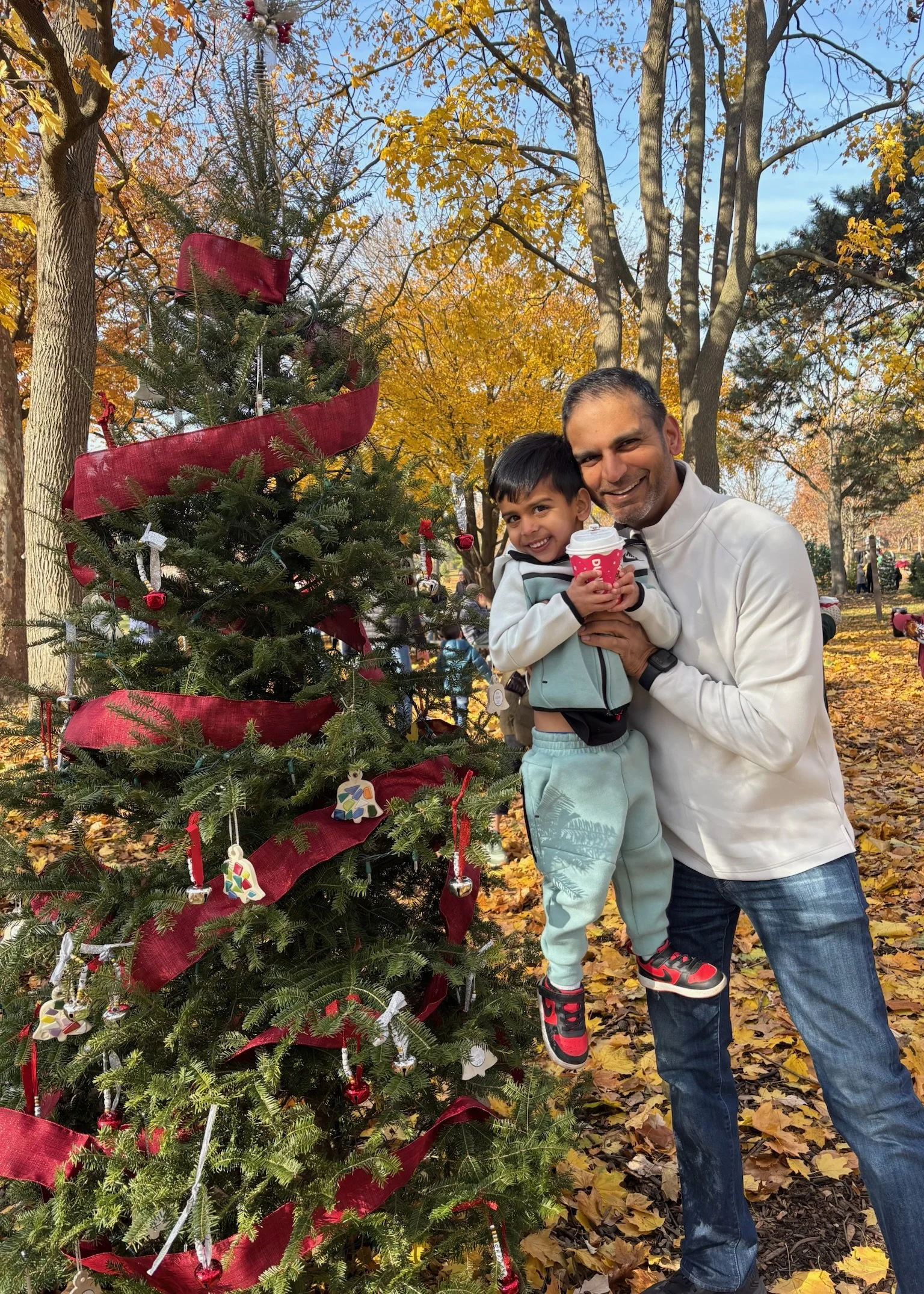 A man and a young boy smiling and holding a cup, standing next to a decorated Christmas tree outdoors.