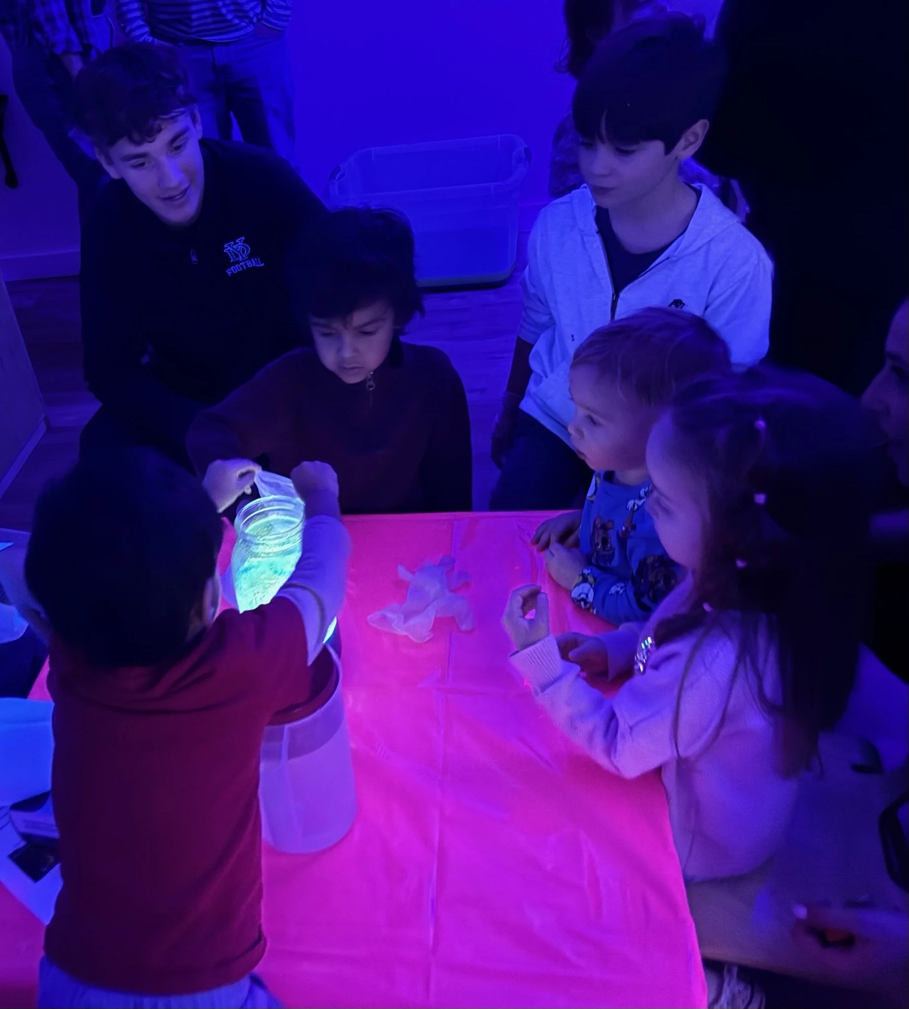 A group of children gathered around a table with a glowing, colorful light, in a dark room with purple and blue lighting, appears to be exploring or observing something on the table.