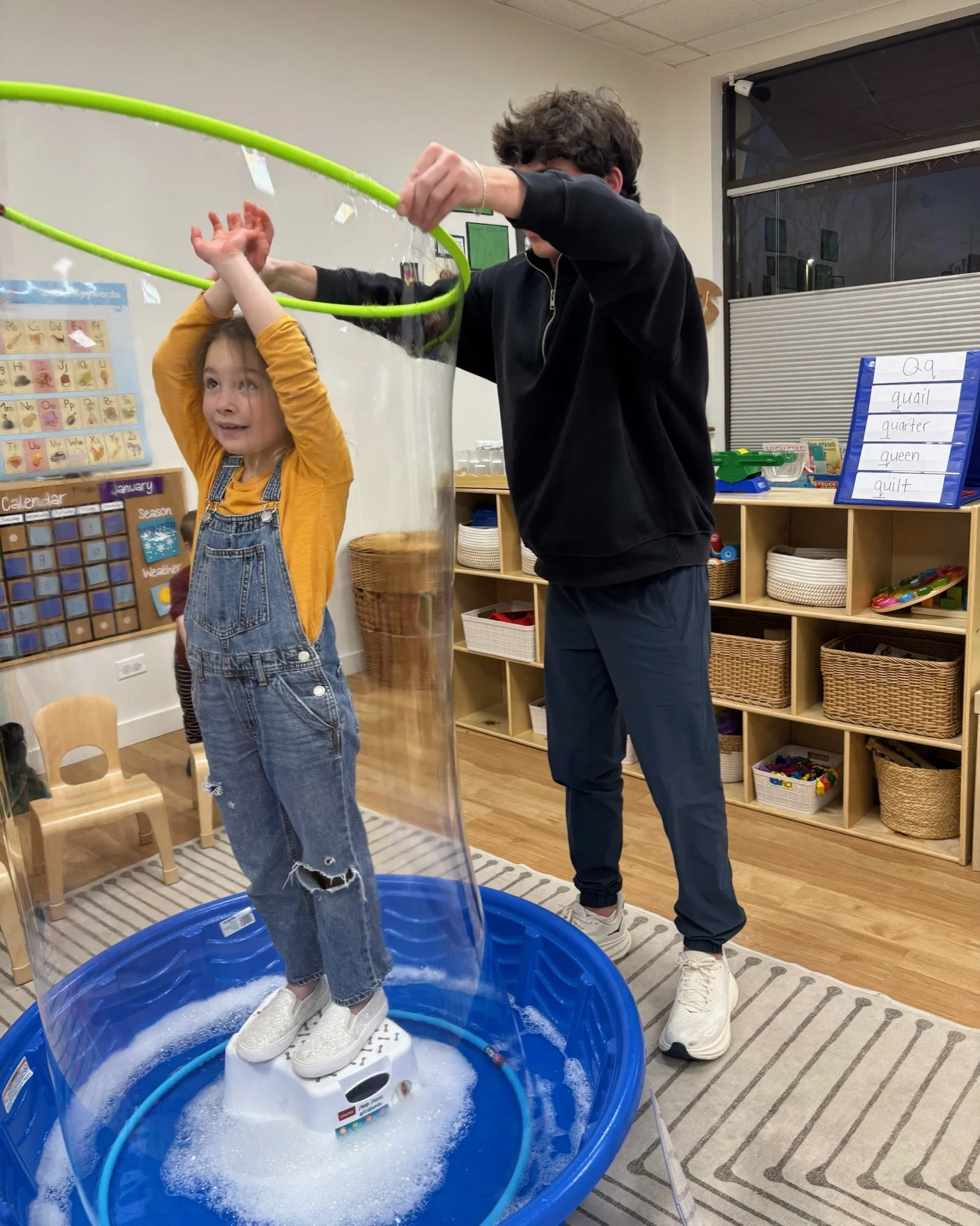 A young girl standing in a small inflatable pool filled with water and bubbles, with a male adult helping her, in a classroom setting with educational posters and shelves in the background.