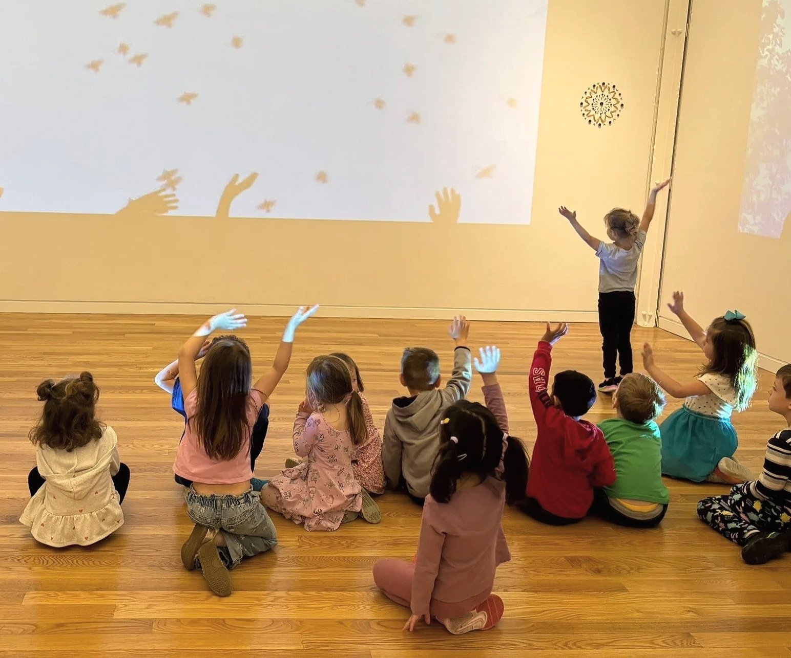 A group of children sitting on a wooden floor in front of a large screen, some raising their hands. One child is standing and gesturing towards the screen, where their shadows are projected as hands and floating shapes.