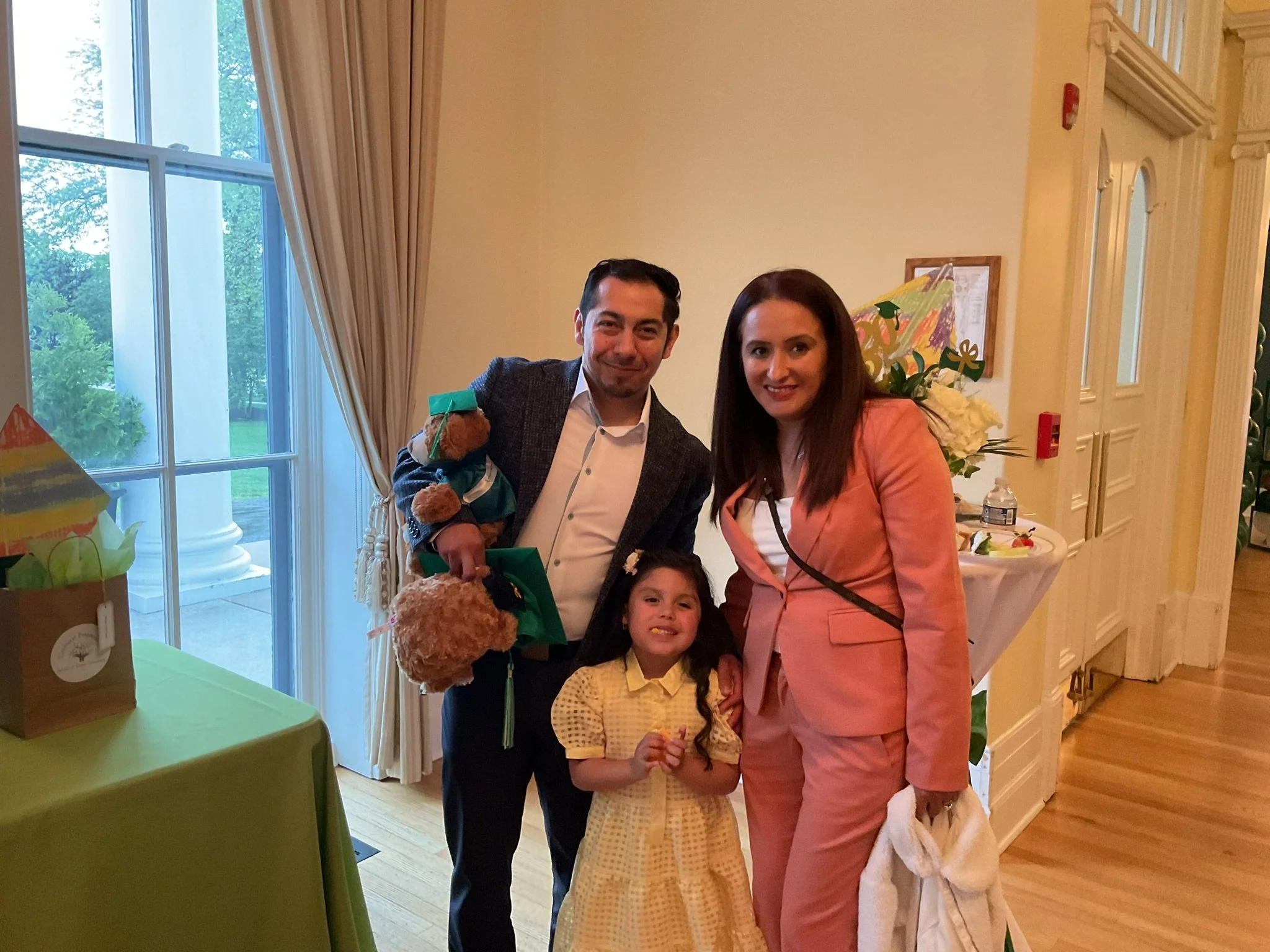 Two children and two adults posing for a picture indoors, near a large window with a view of trees outside, at a celebration or event, decorated with flowers and balloons.