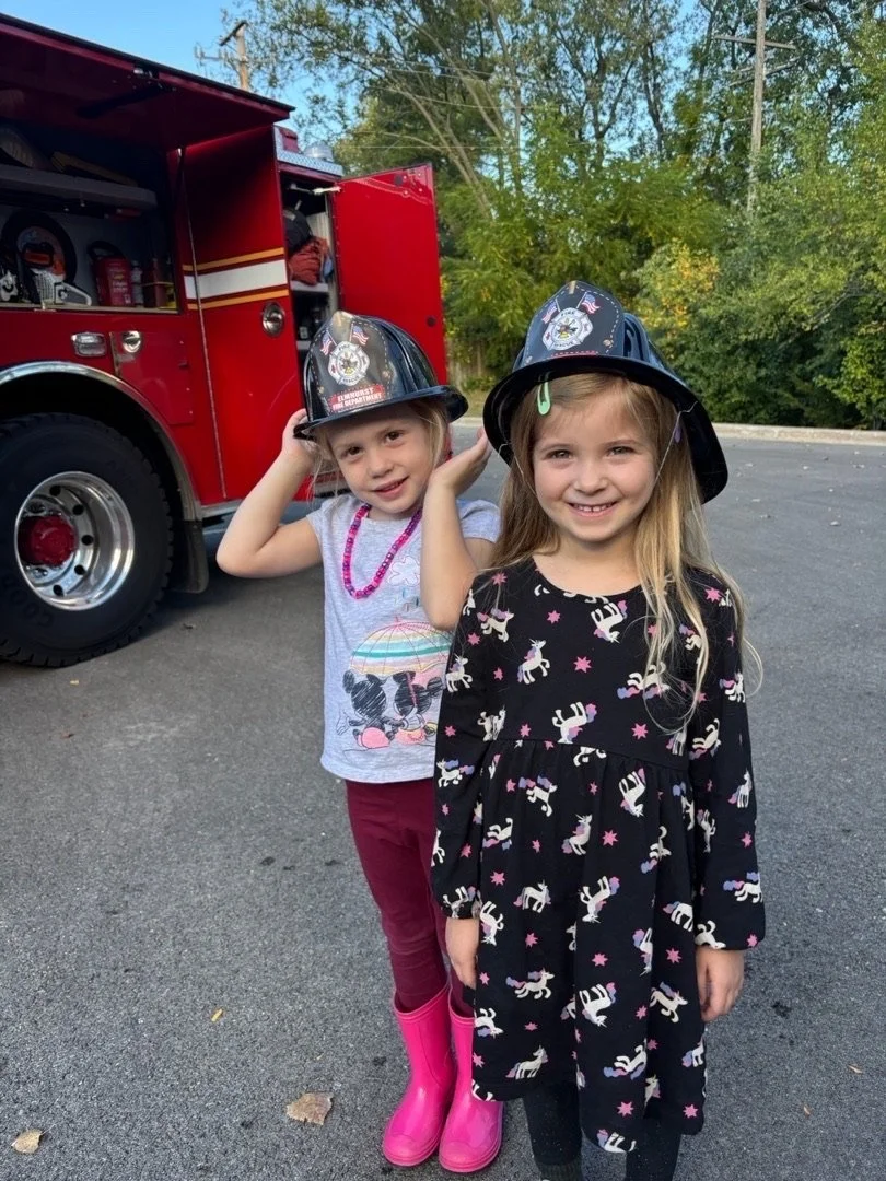 Two young girls standing outdoors next to a red fire truck, wearing firefighter helmets.