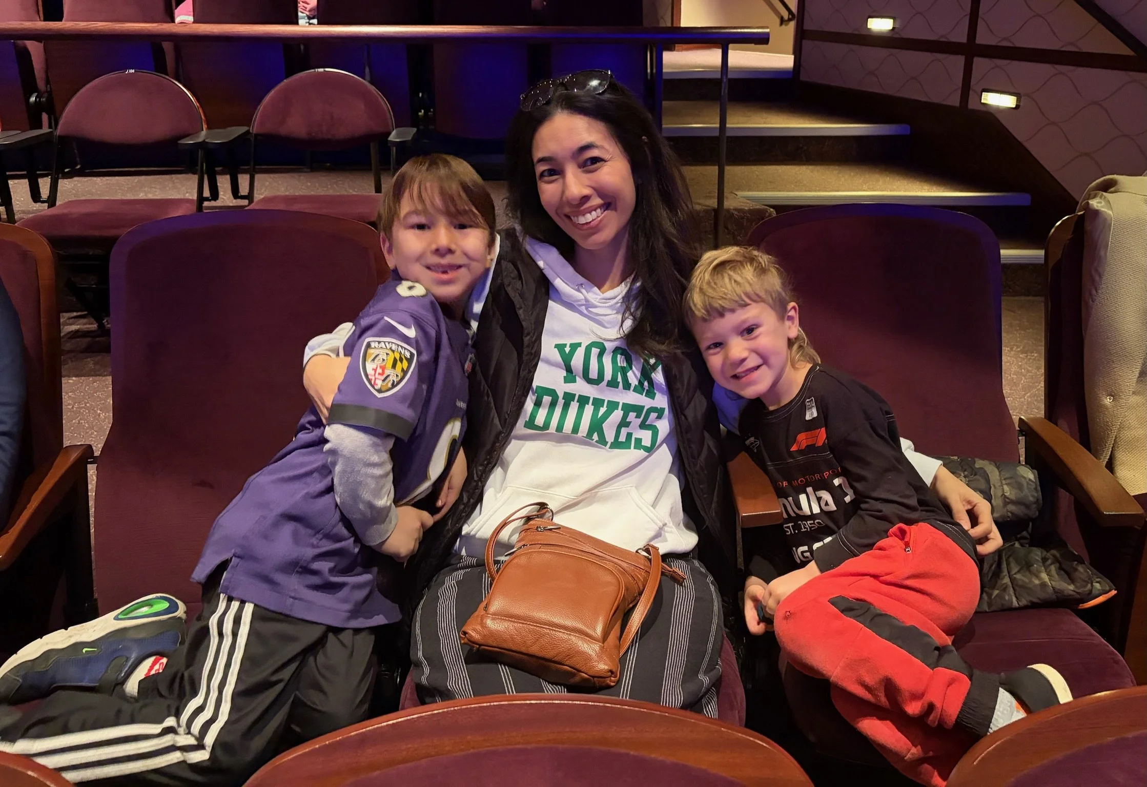 A woman and two young boys sitting in theater seats, smiling at the camera.