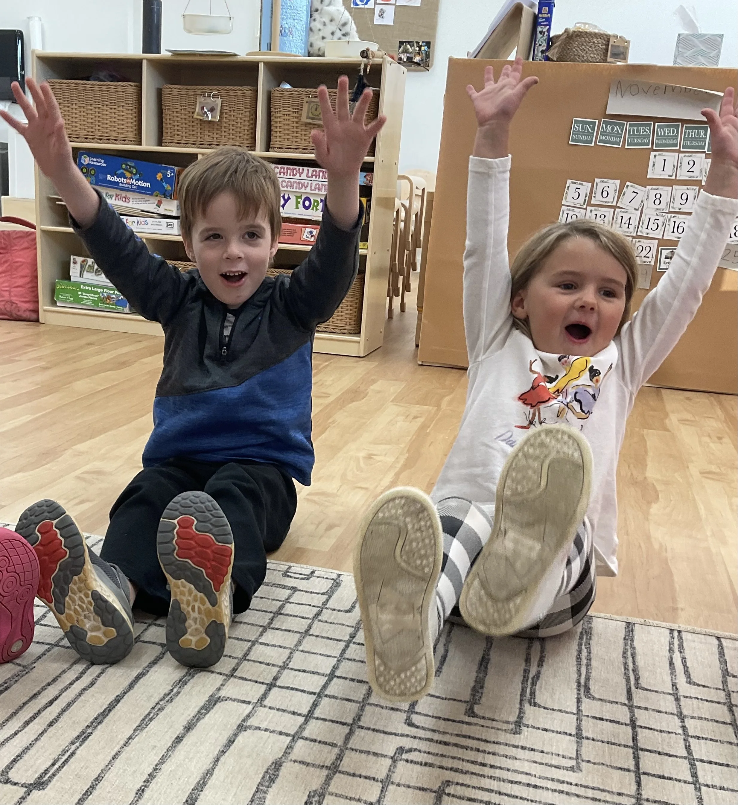 Two children sitting on the floor in a classroom with their legs extended and feet towards the camera, raising their hands with open mouths, appearing excited. The classroom has wooden flooring, a patterned rug, and shelves with toys and educational materials in the background.