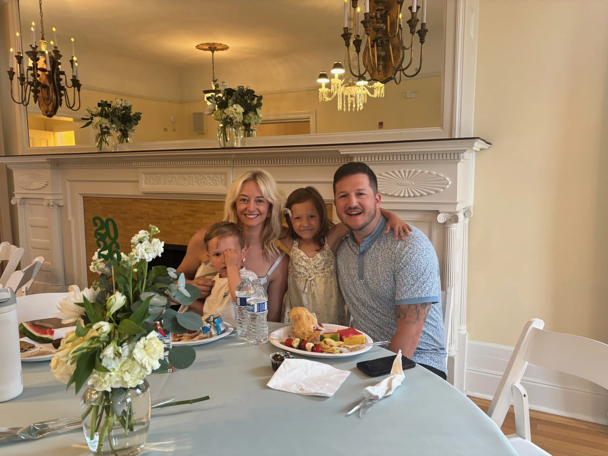 A family of five celebrating a special occasion in a decorated room, sitting at a table with various desserts and floral arrangements, with candles and chandeliers hanging from the ceiling.