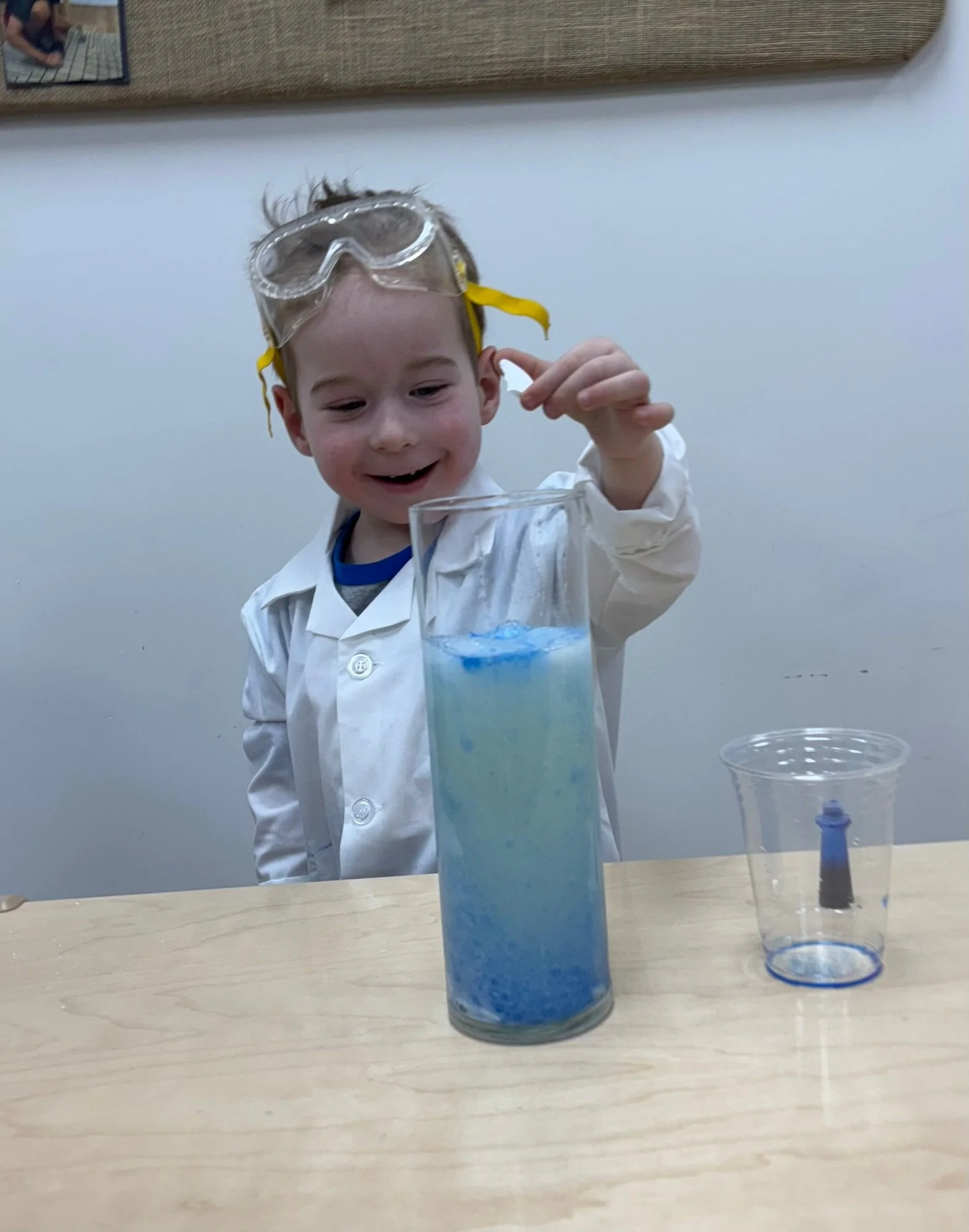 A young boy wearing goggles and a white lab coat is conducting a science experiment. He is holding a small container and smiling, with a tall glass filled with a blue mixture and a smaller container with a dark substance on a table.
