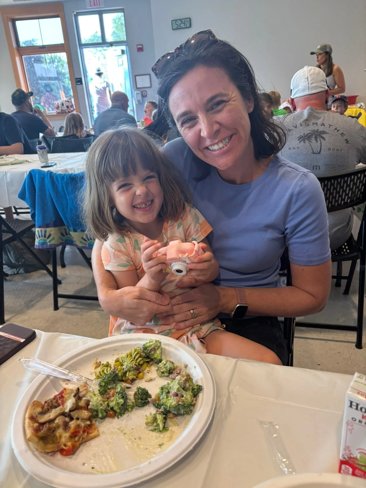 A woman and a young girl smiling and sitting together at a table during a meal in a restaurant or cafe. The table has a white disposable plate with remnants of pizza and salad. In the background, there are other people dining, a person standing near 