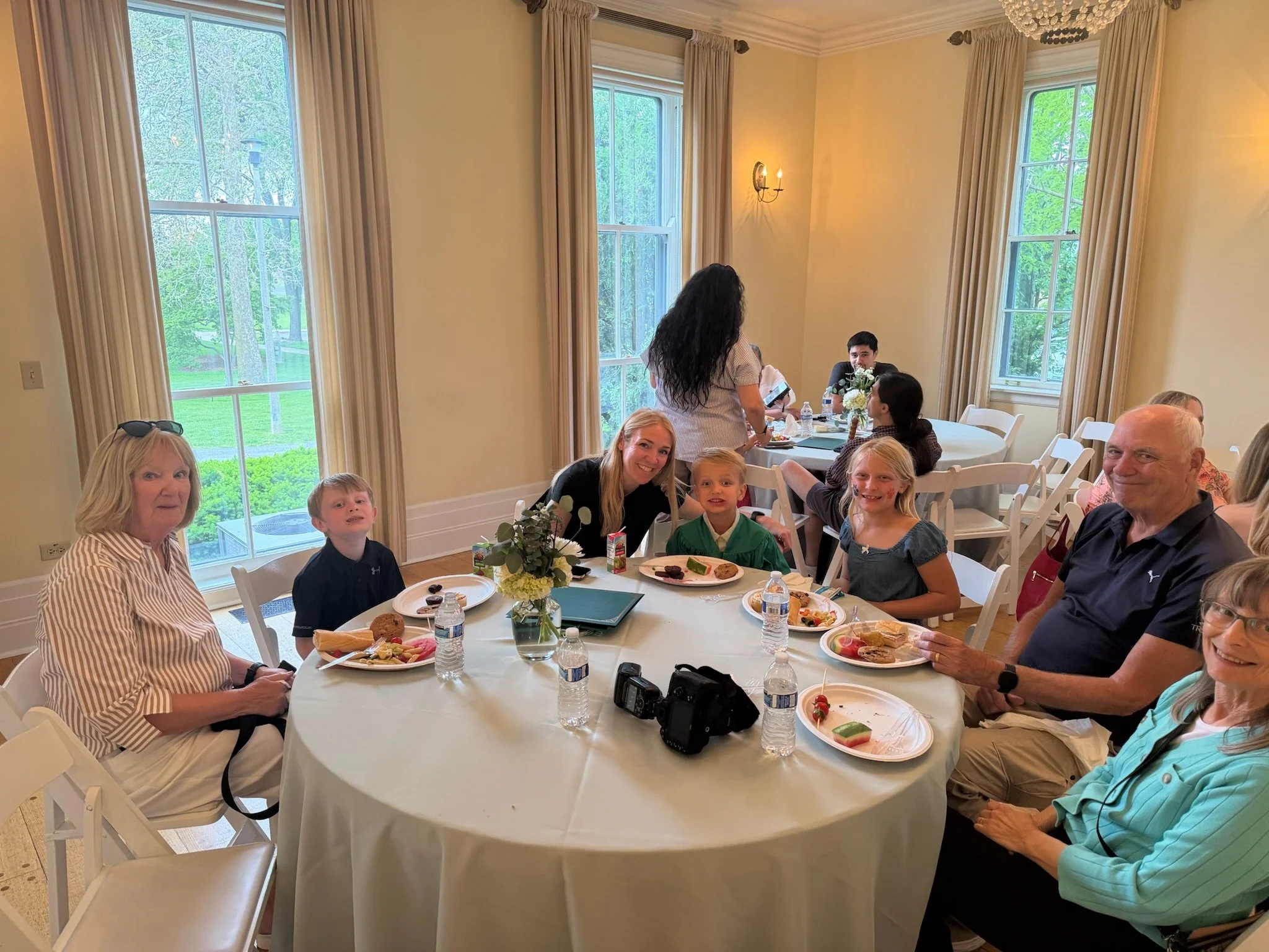 Family and friends gathered around a table at a celebration, enjoying food and drinks in a bright room with large windows showing outdoor trees.