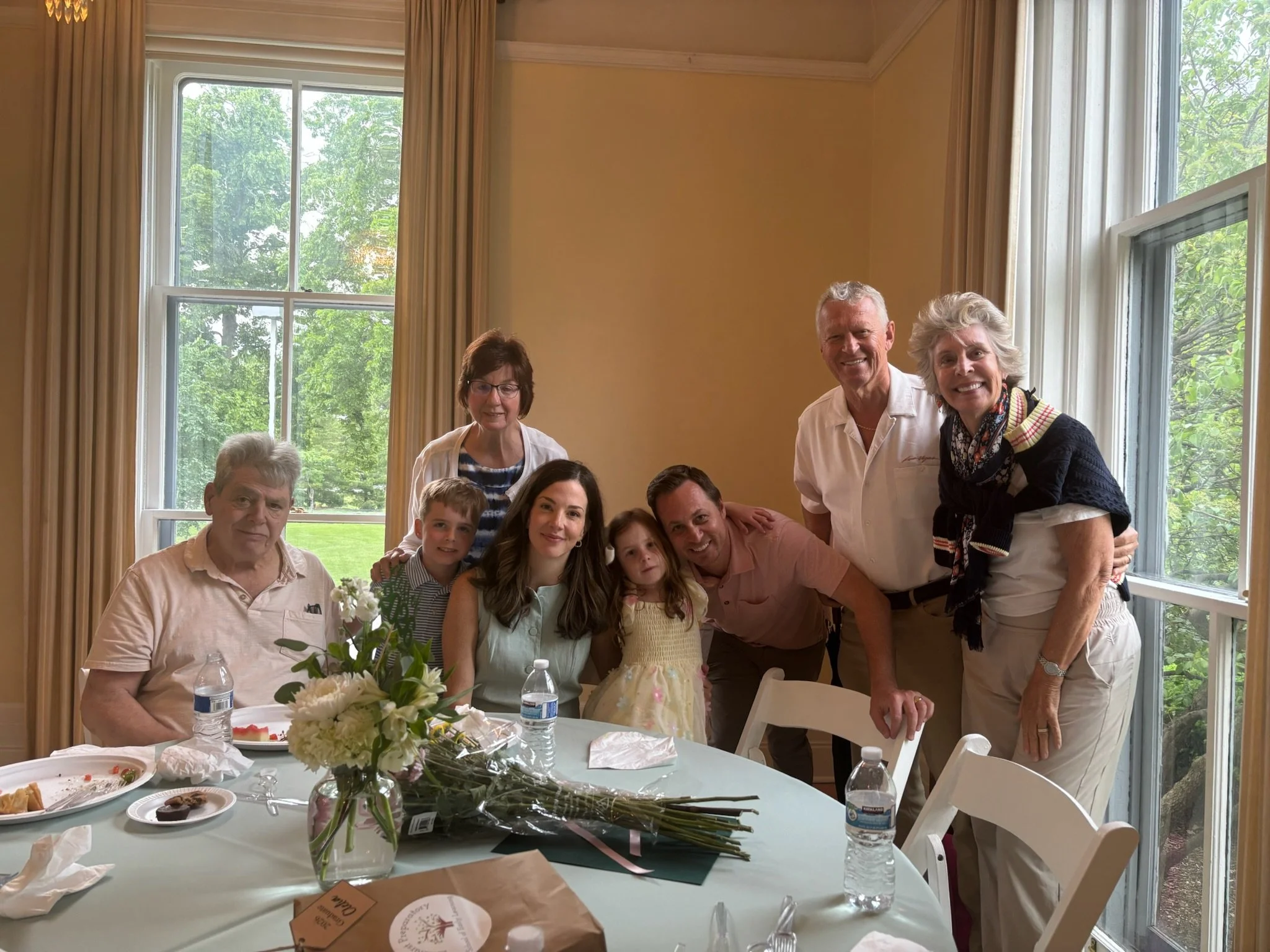 A group of family and friends gathered around a table inside a bright room with large windows, some of them standing and smiling at the camera, with flowers, food, and water bottles on the table.