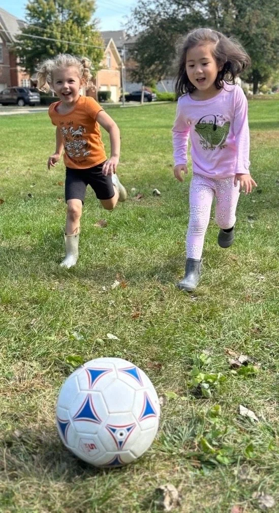 Two young girls running towards a soccer ball on a grassy field, smiling and enjoying outdoor play.