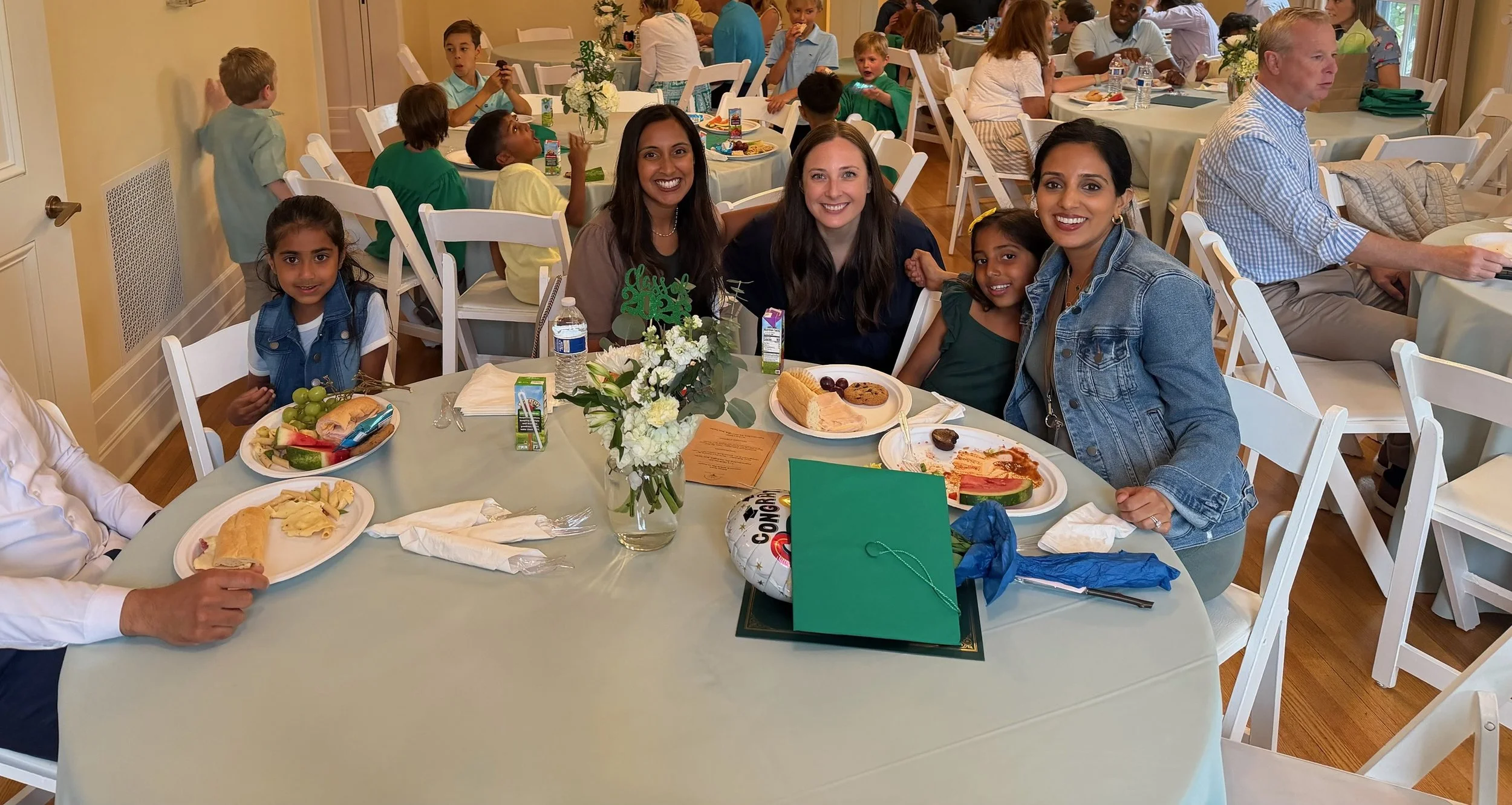 A group of people, including children and adults, sitting around a table at a celebratory gathering or party. The table is decorated with a floral centerpiece and has plates of food, drinks, and party favors. In the background, other tables with gues