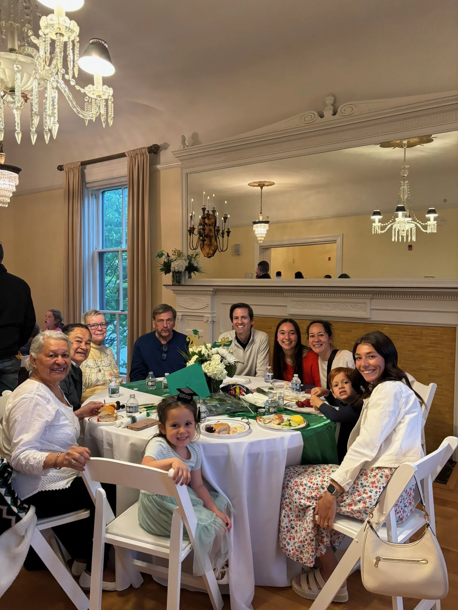 A group of people seated around a round table at a celebration, with a mix of children and adults, in a decorated indoor setting with chandeliers and a mirror on the wall.