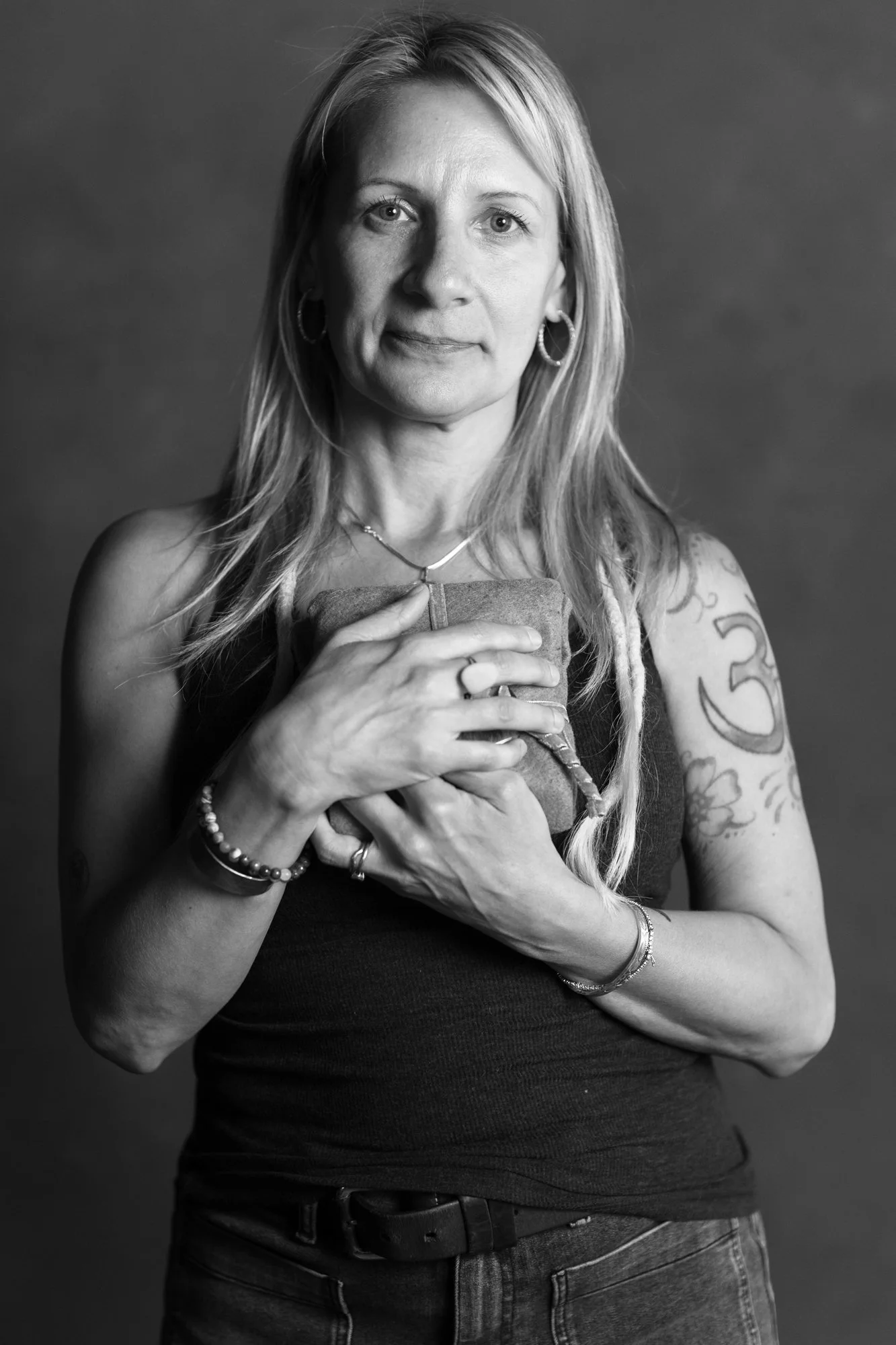 A black and white portrait of a woman with long blonde hair, wearing hoop earrings, a necklace, a black sleeveless top, and jeans. She is holding her top with both hands and has tattoos on her left arm.
