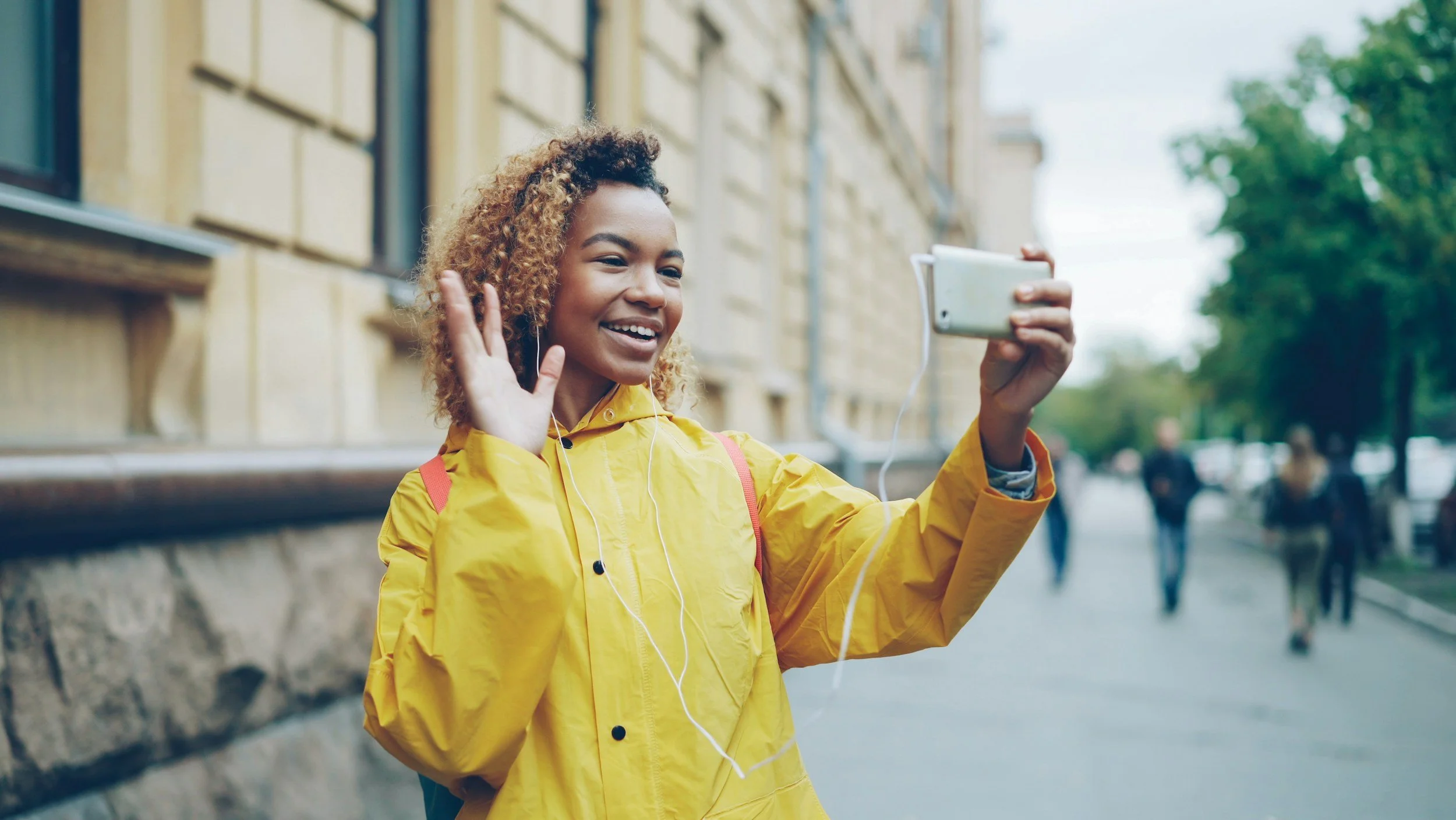 Woman in a yellow jacket smiling and waving at a smartphone on a city street