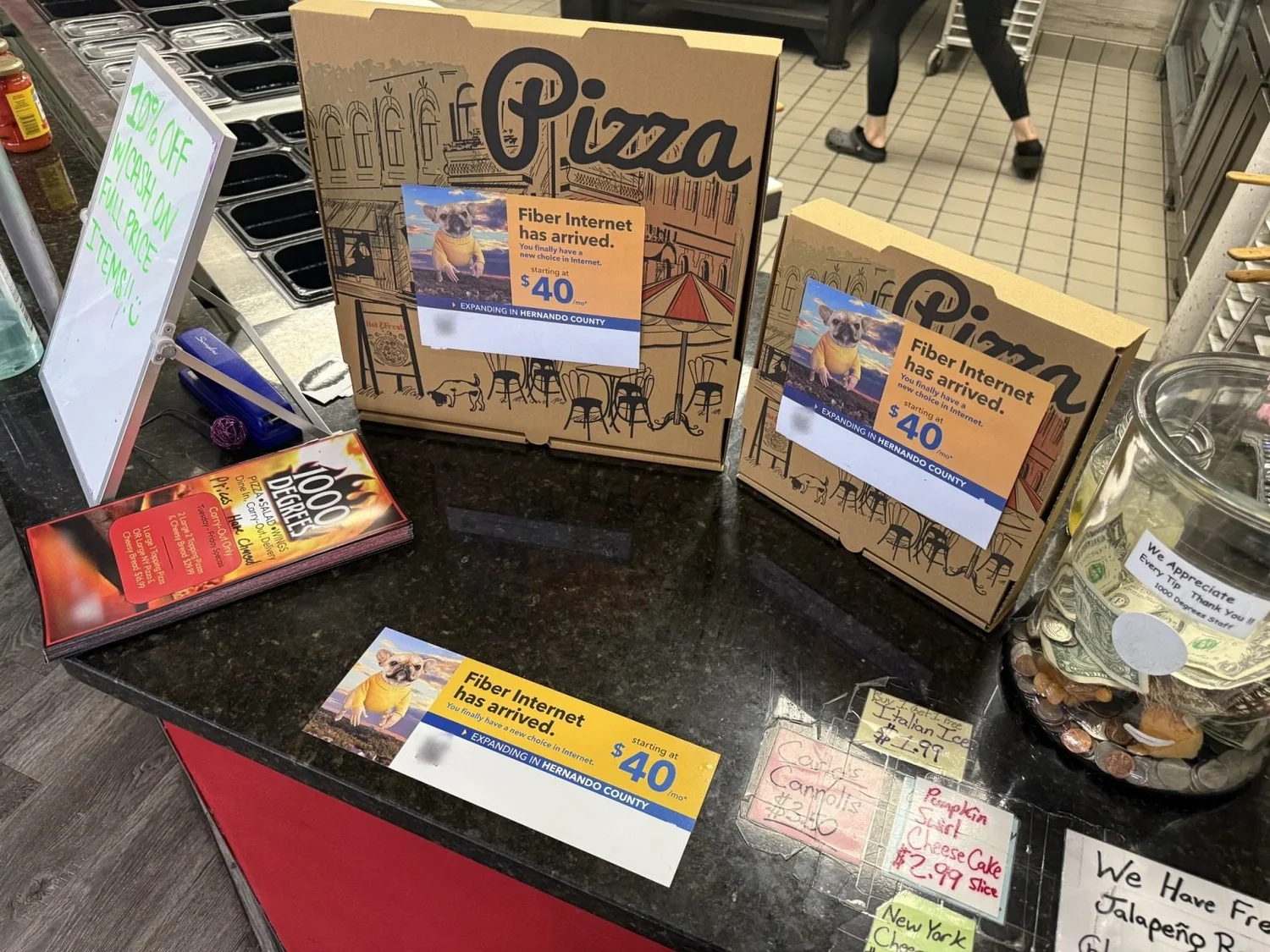 Pizza box toppers on stacked pizza boxes beside a counter card at a pizzeria register