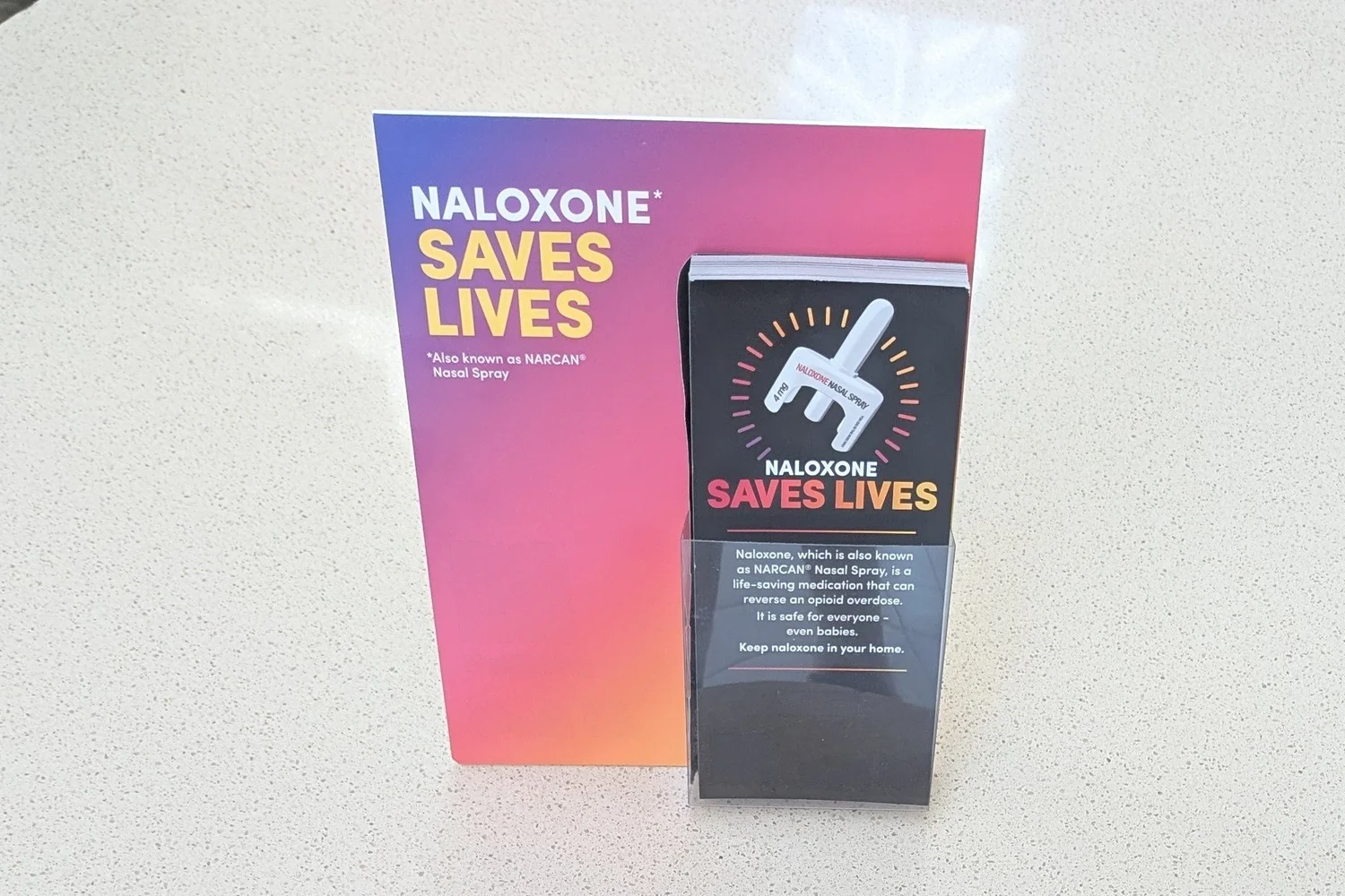 Naloxone take-one cards in an acrylic holder in front of a matching sign on a clinic waiting-area table