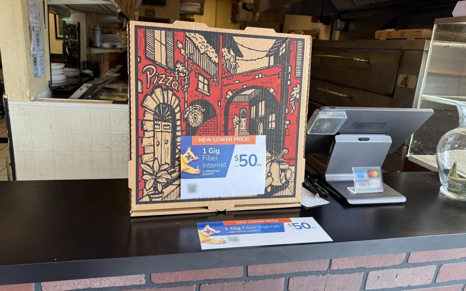 Illustrated pizza box with a topper, next to a counter card and card reader on a pizzeria counter