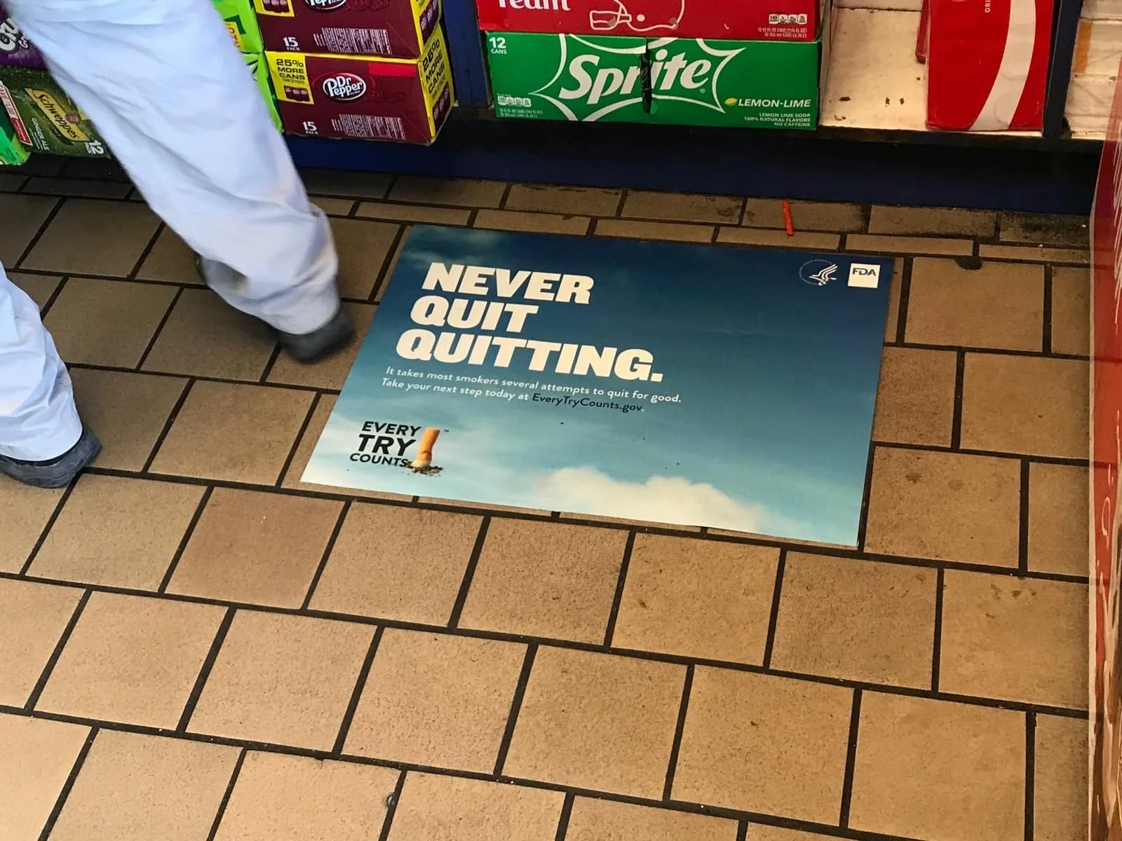 Convenience store advertising - Floor with an anti-smoking poster near beverage boxes, displaying the text 'Never Quit Quitting' along with encouragement to quit smoking from the FDA.