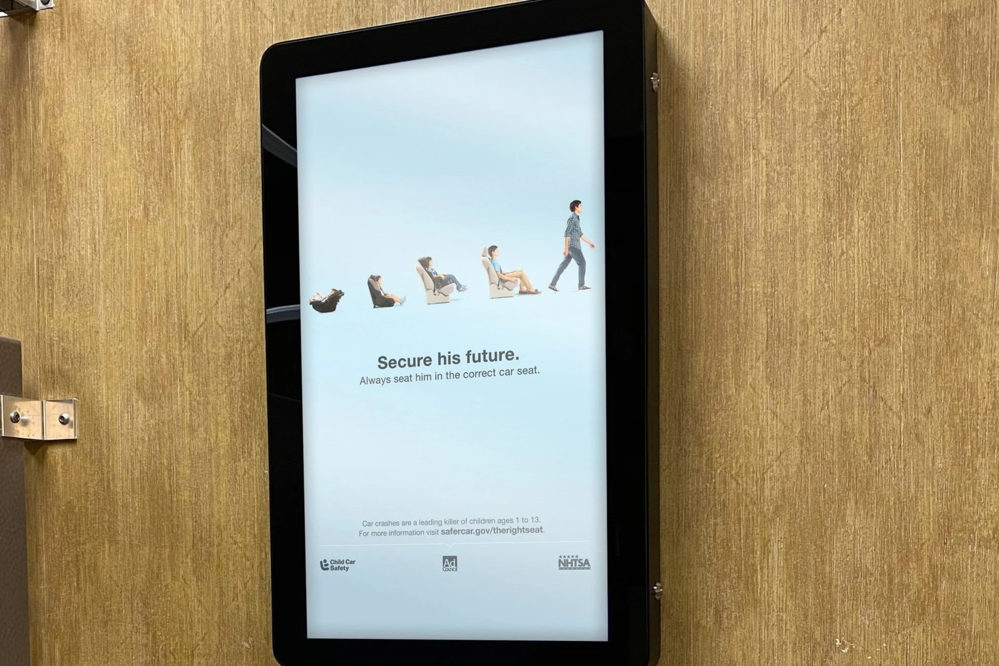 Digital sign showing the progression of a person sitting in various car seats to walking, with the message 'Secure his future. Always seat him in the correct car seat.' and a note referencing child car safety.