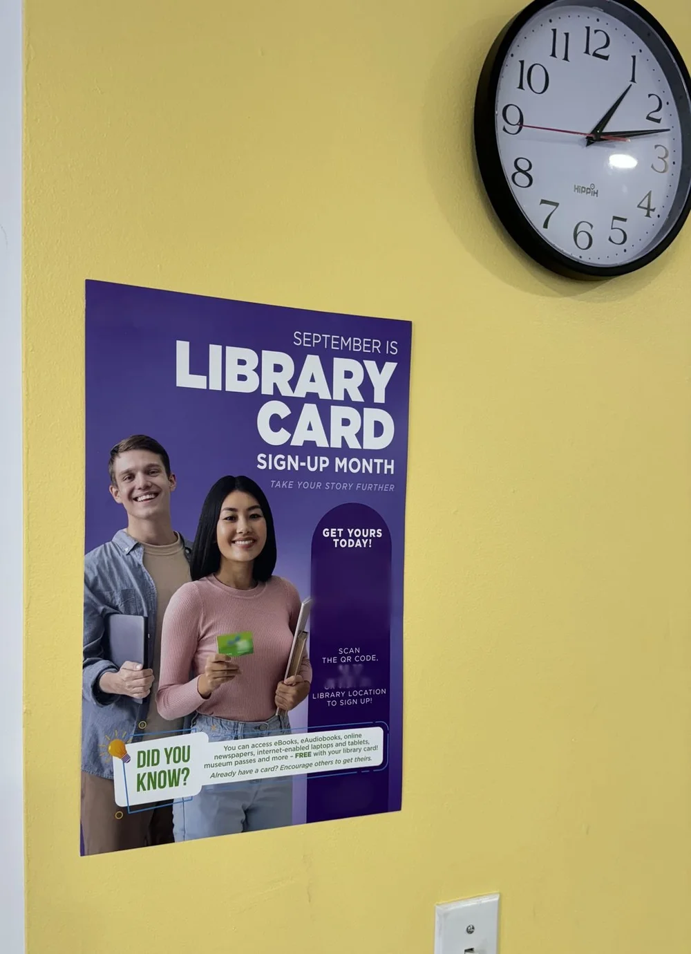 Library card sign-up poster on a yellow school hallway wall beside a clock