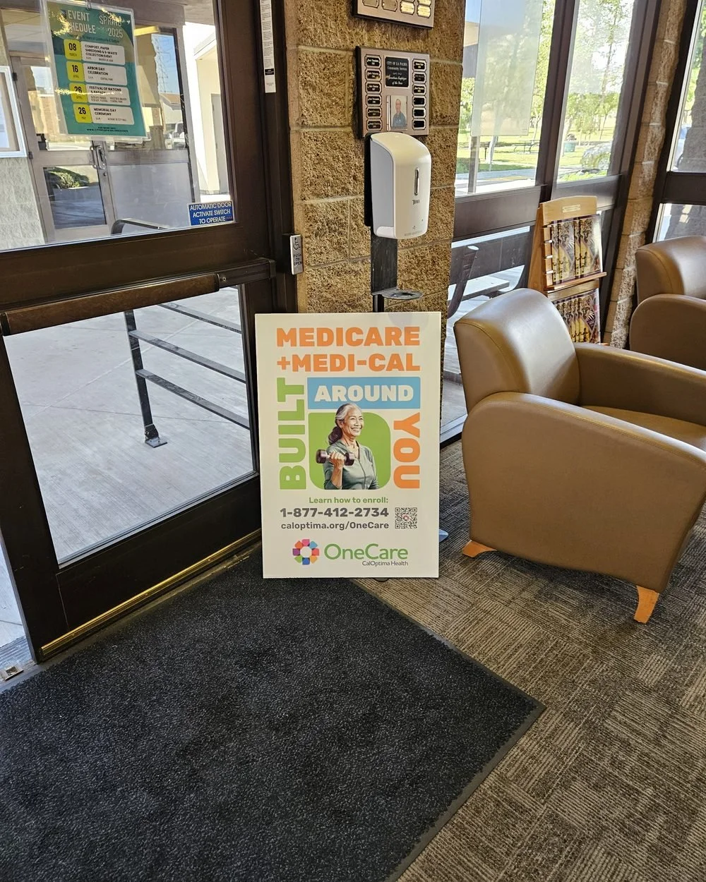Medicare and Medi-Cal health plan enrollment poster standing on the floor inside a community center entrance, beside lobby armchairs